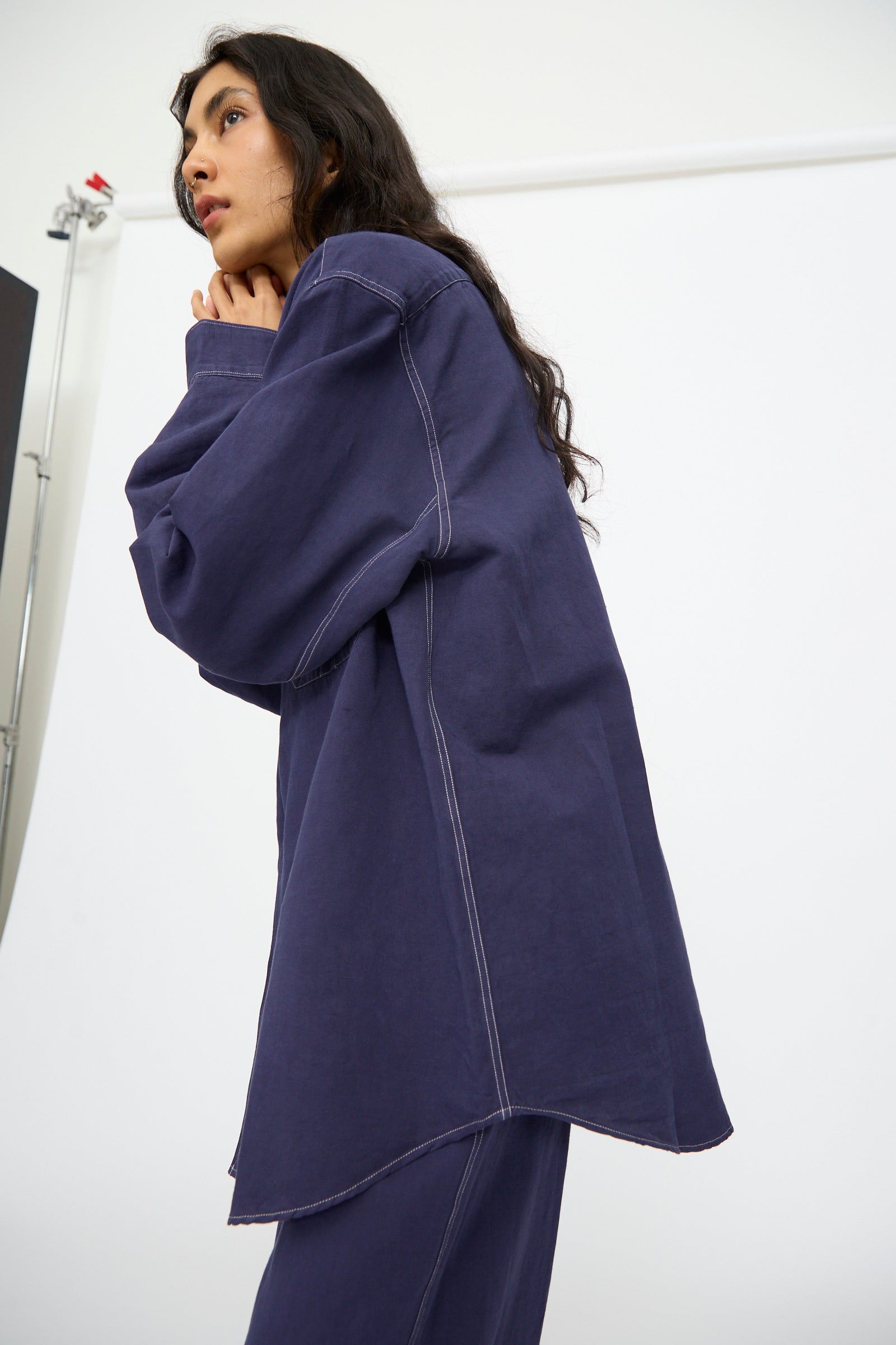 A woman models the Studio Nicholson Louro Utility Pocket Shirt in Indigo, styled with matching pants. She stands against a white backdrop, gazing sideways with her hands near her face.