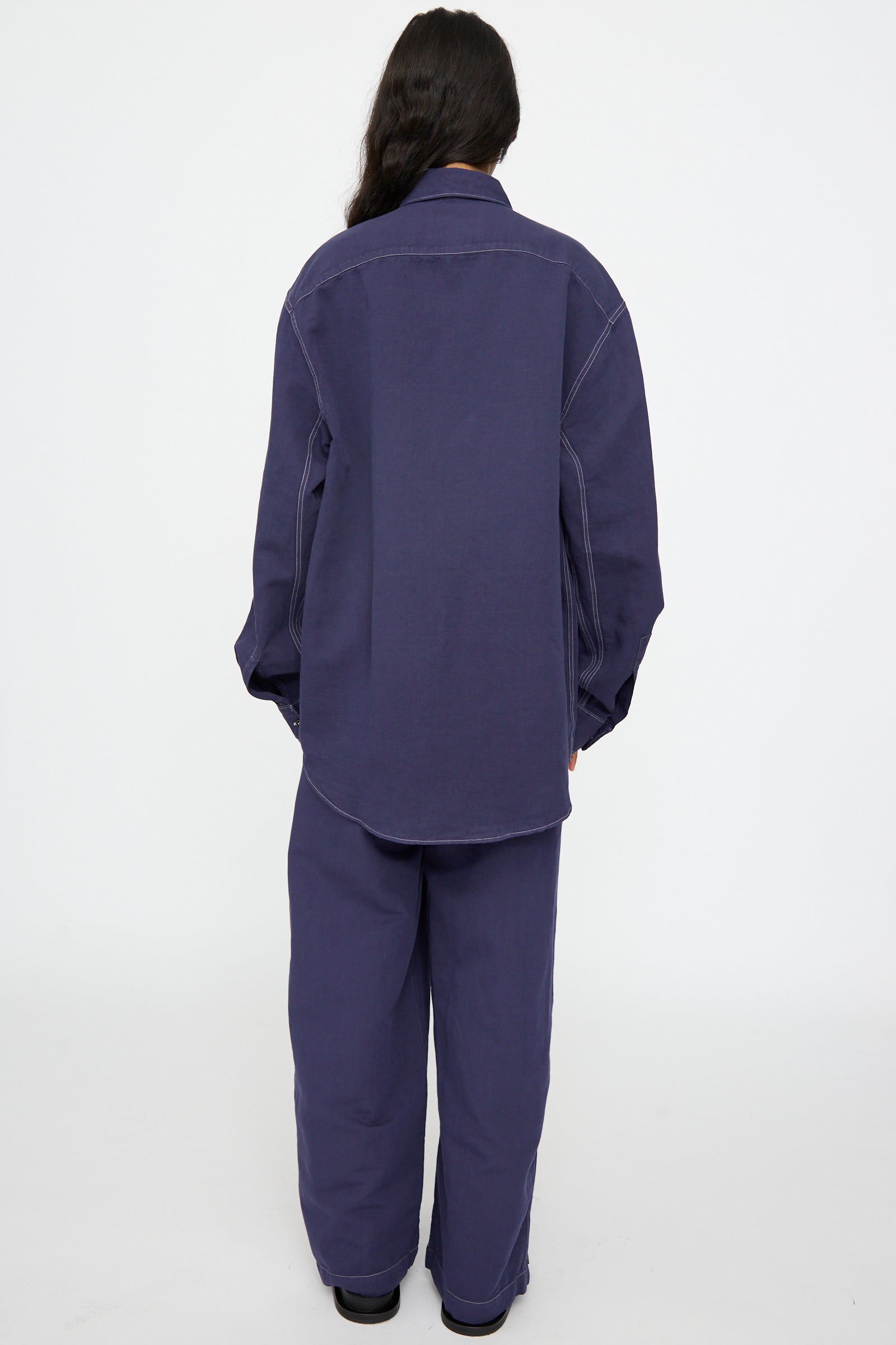 A person stands with their back to the camera, wearing the Studio Nicholson Louro Utility Pocket Shirt in Indigo and matching pants against a plain white background.