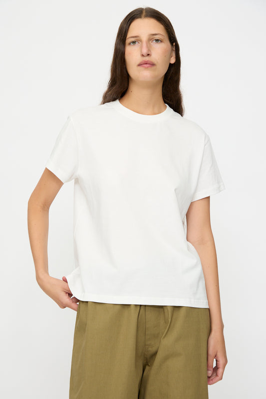 A person with long brown hair wears the Studio Nicholson Marine Perfect Tee in Off White, featuring a boxy fit and short sleeves, paired with olive green pants, standing against a plain white background.