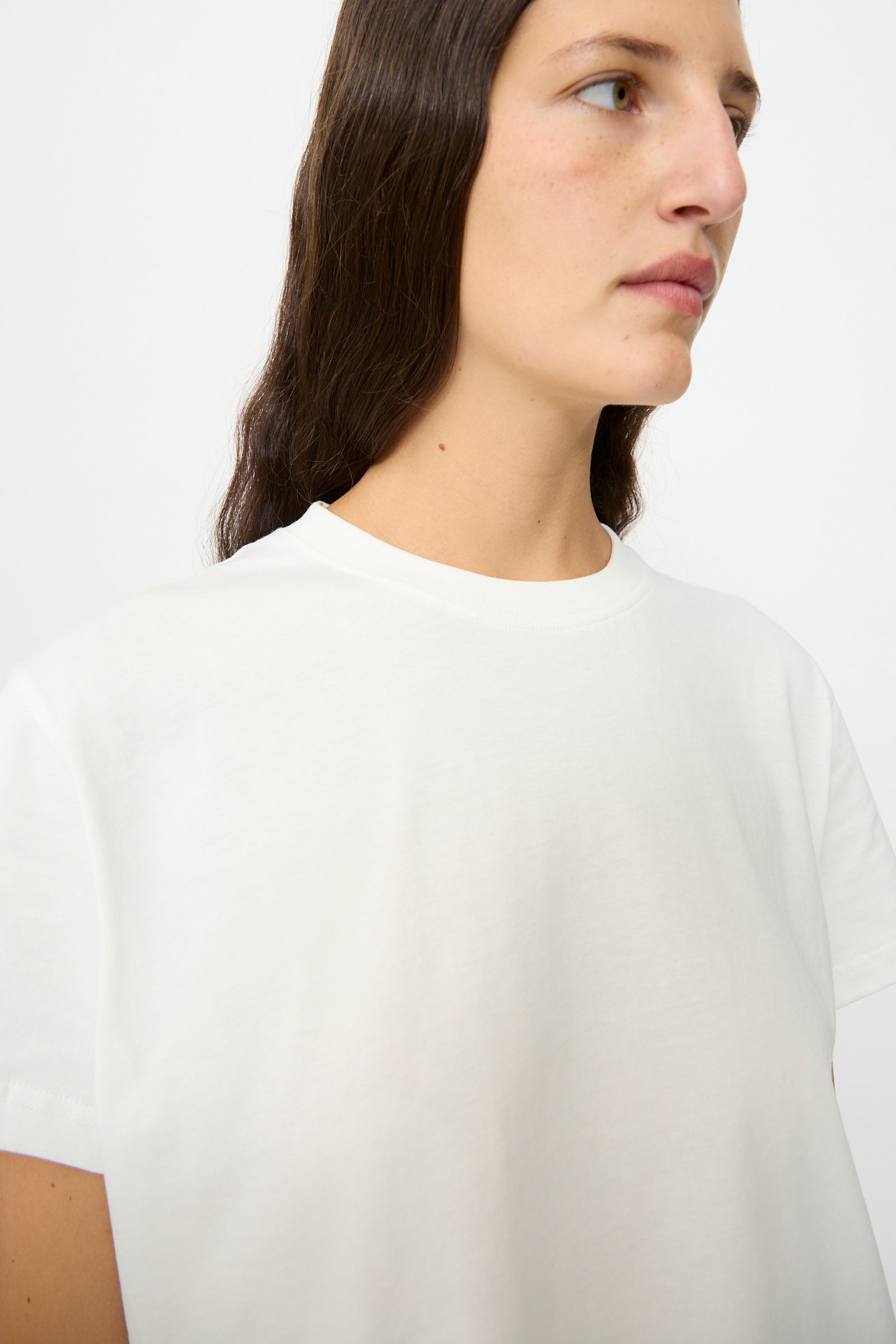 A woman with long brown hair wears the Studio Nicholson Marine Perfect Tee in Off White, a classic cotton jersey short sleeve top, shown from the shoulders up against a plain background.