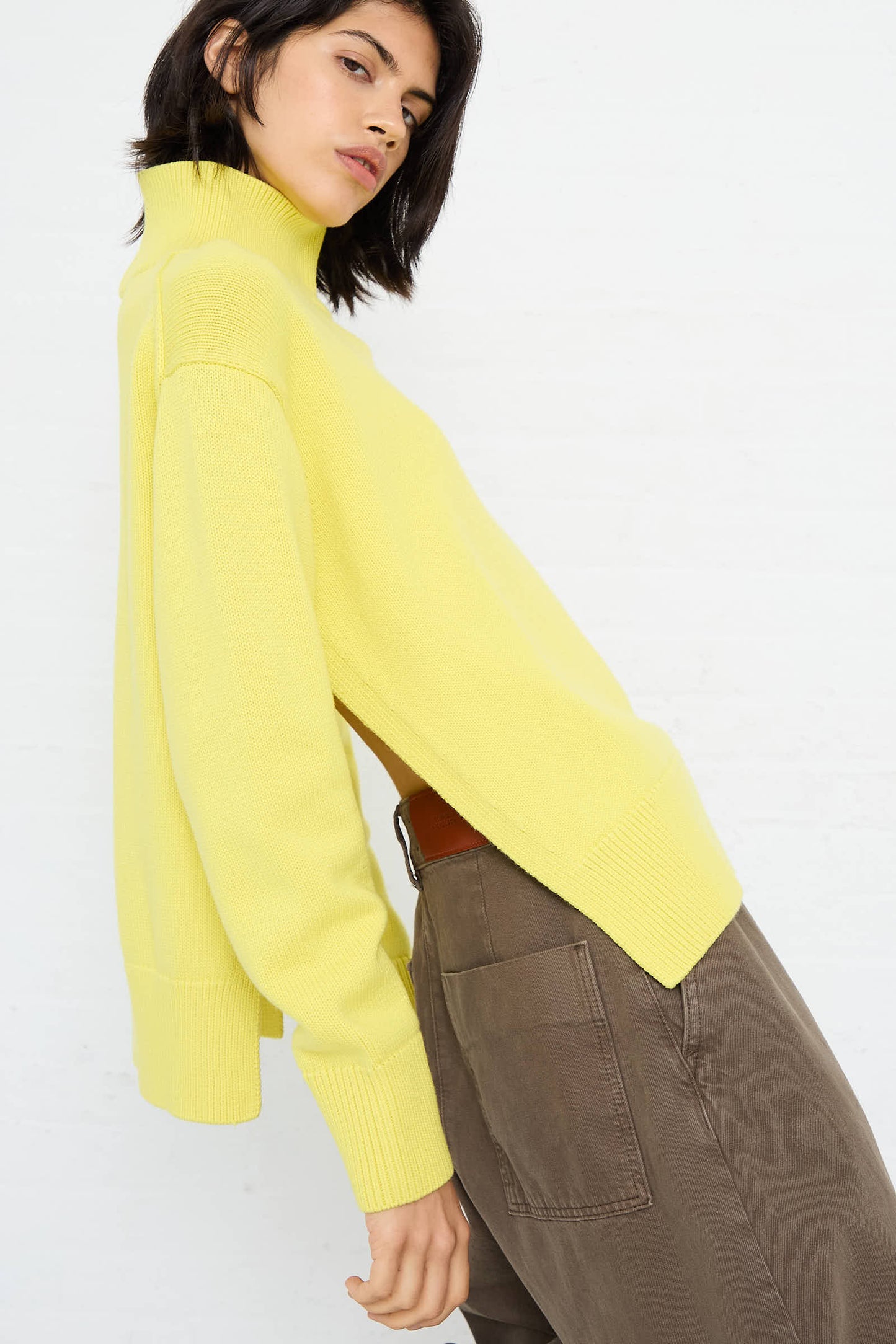 A person wearing an oversized Merino Cotton Patria Roll Neck Sweater in Acid Yellow from Studio Nicholson and brown pants, posing against a white background.