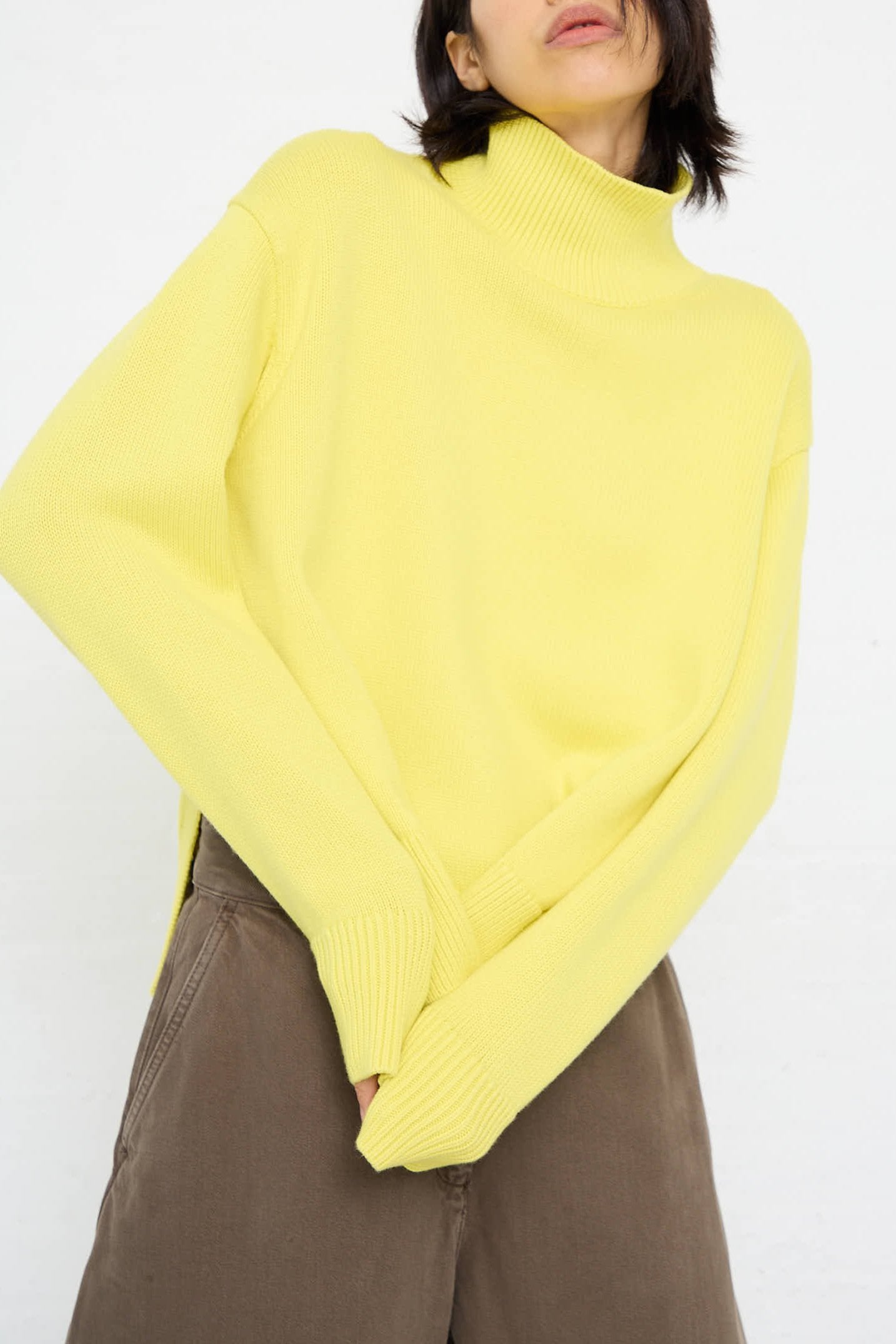 A person with medium-length dark hair wearing the Studio Nicholson Merino Cotton Patria Roll Neck Sweater in Acid Yellow and brown pants stands against a plain background.