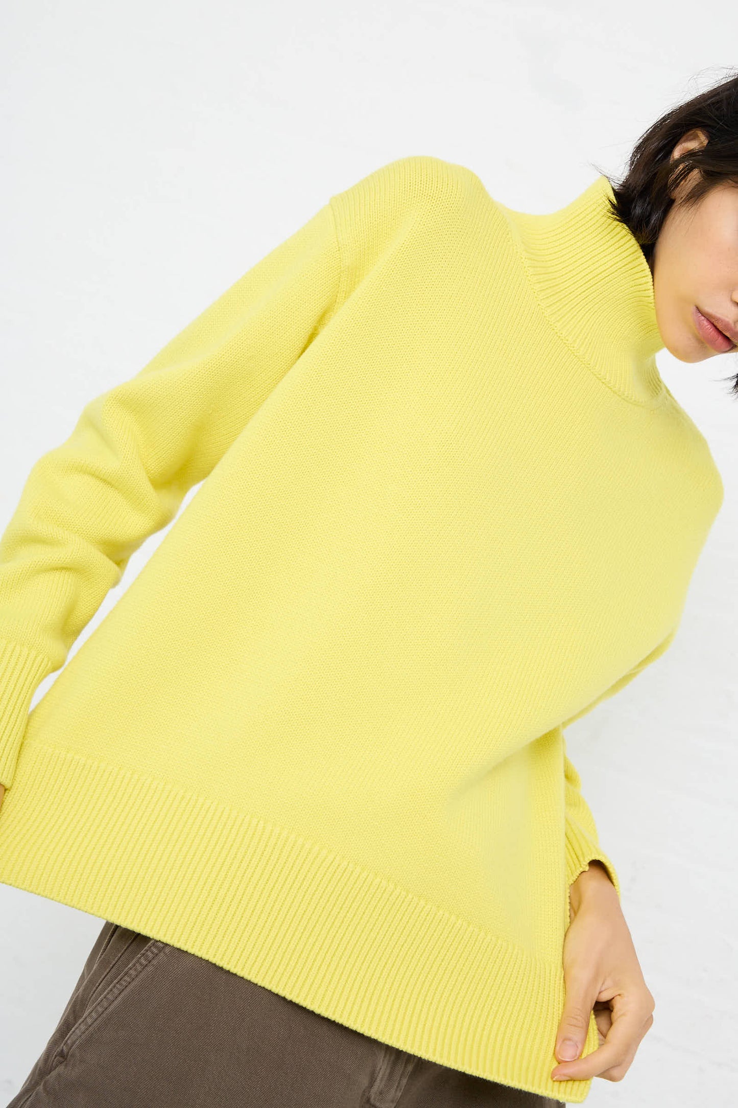 A person wearing Studio Nicholson's Merino Cotton Patria Roll Neck Sweater in Acid Yellow and brown pants, partially cut off from the image at the head and legs, stands against a white background.