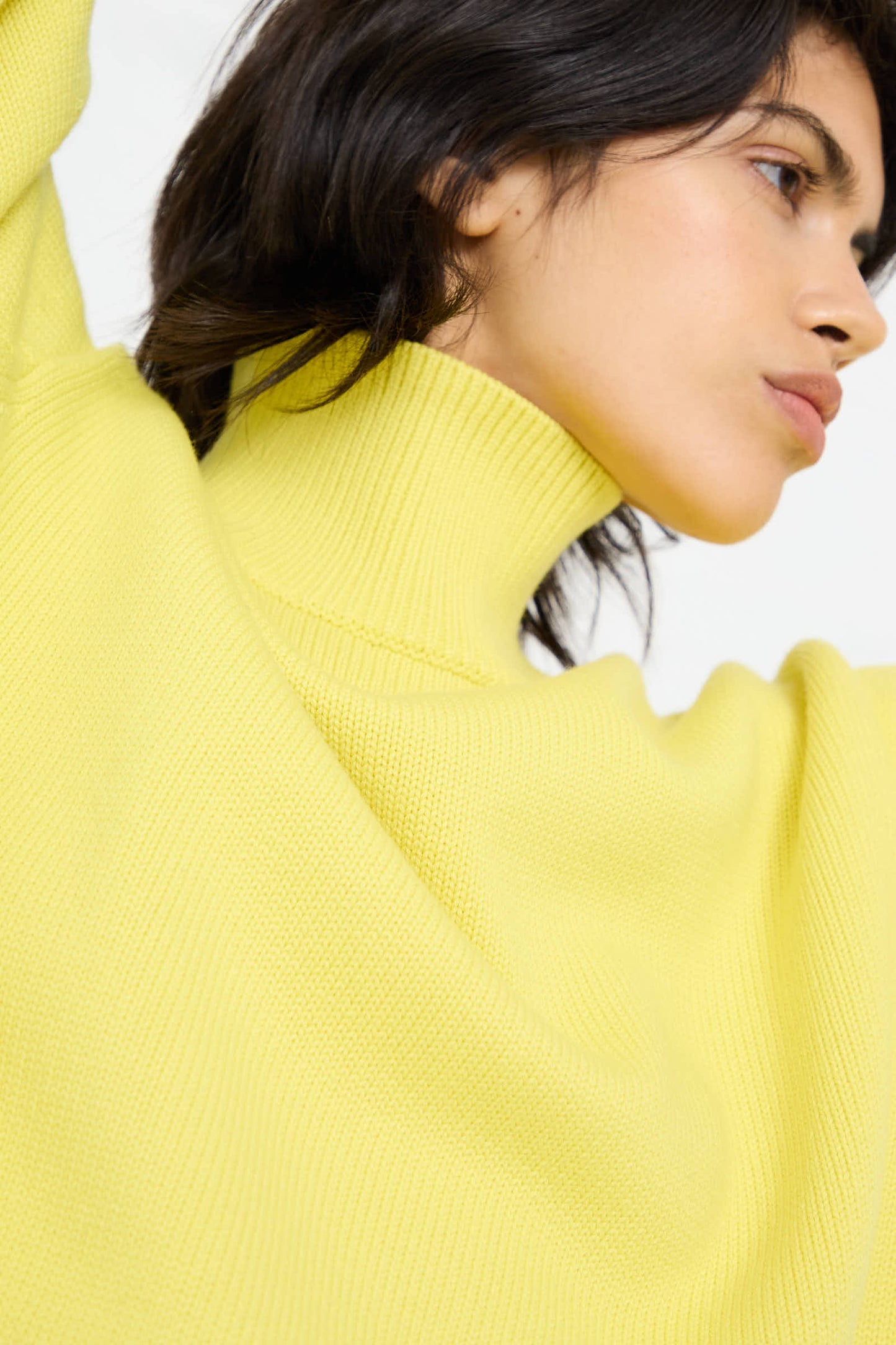 Close-up of a person with dark hair wearing the Merino Cotton Patria Roll Neck Sweater in Acid Yellow by Studio Nicholson. The person's face is turned slightly to the side.