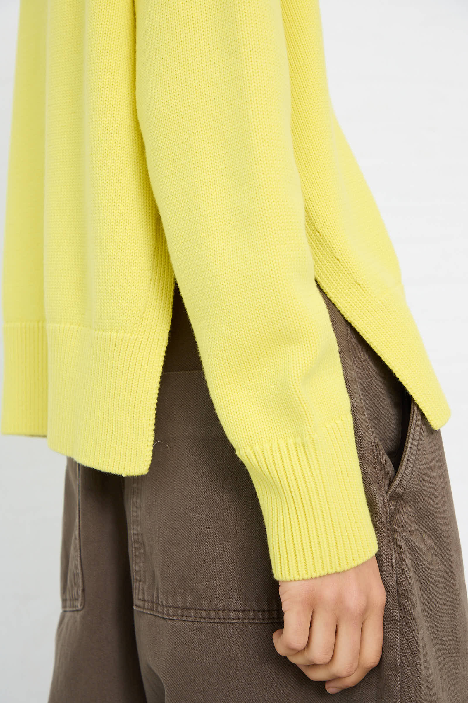 A person wears the Studio Nicholson Merino Cotton Patria Roll Neck Sweater in Acid Yellow with brown pants. Only the lower half of the relaxed fit sweater, featuring side slits, and pants are visible. The left hand is partially tucked into the pocket.