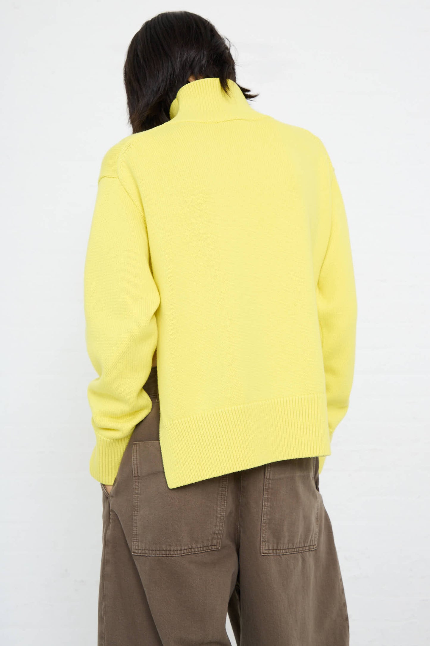 An individual, seen from behind, is wearing a relaxed fit Studio Nicholson Merino Cotton Patria Roll Neck Sweater in Acid Yellow and brown pants with large back pockets, standing against a plain white background.