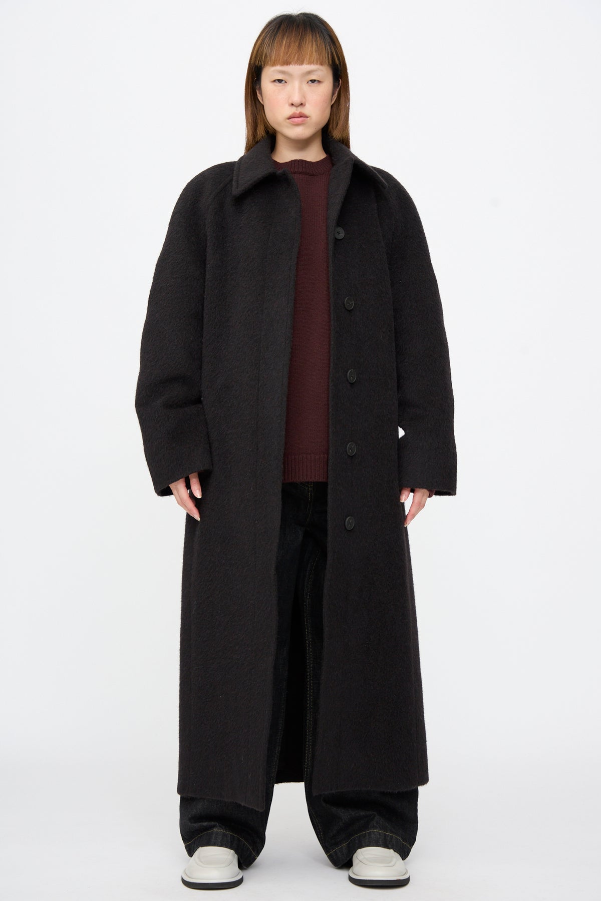 A person wears a long, oversized dark brown Studio Nicholson Mohair Kober Coat in Cola over a burgundy sweater and dark wide-leg trousers.