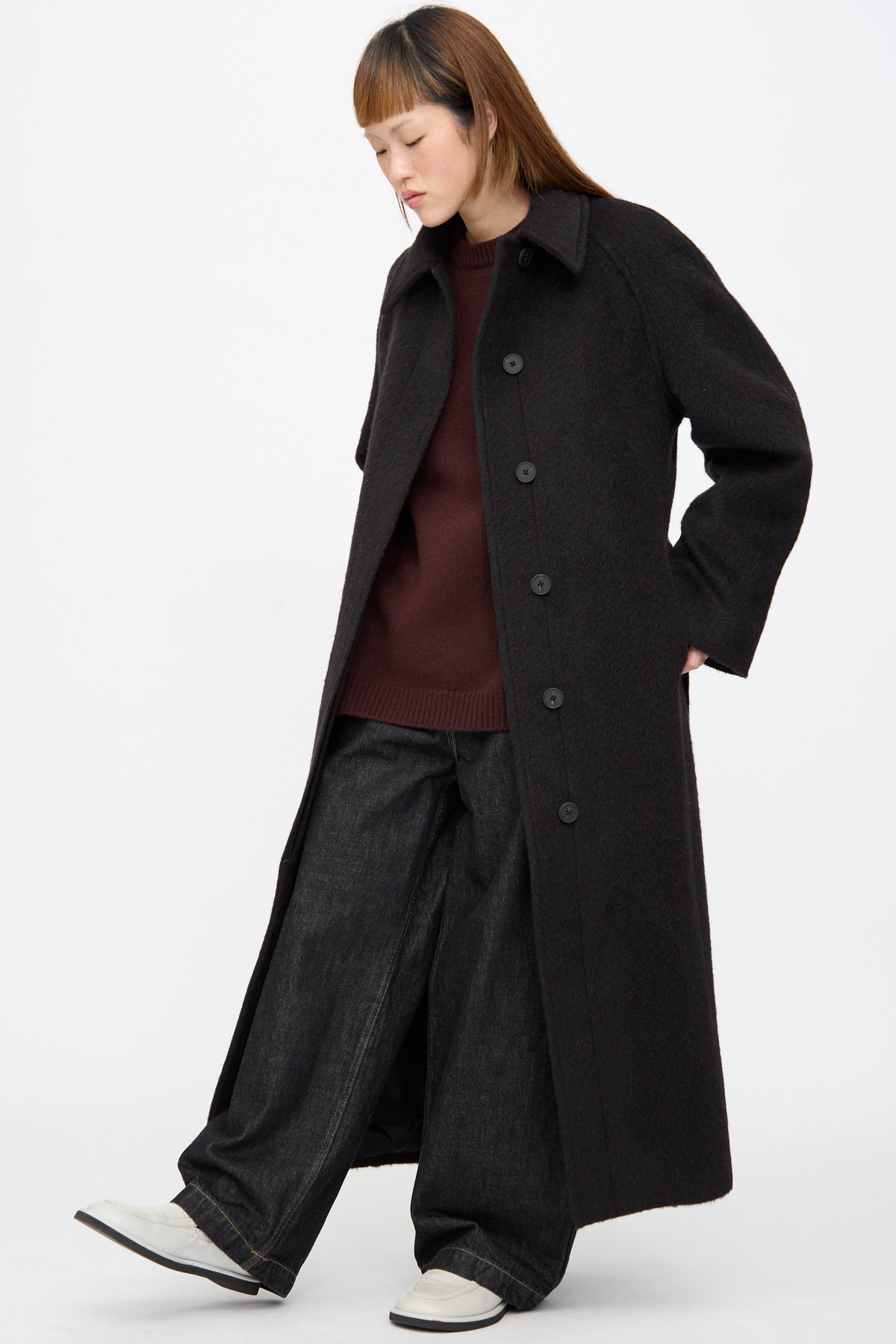 A person wears an oversized, long Studio Nicholson Mohair Kober Coat in Cola over a burgundy sweater and wide-leg dark denim pants, with white loafers.
