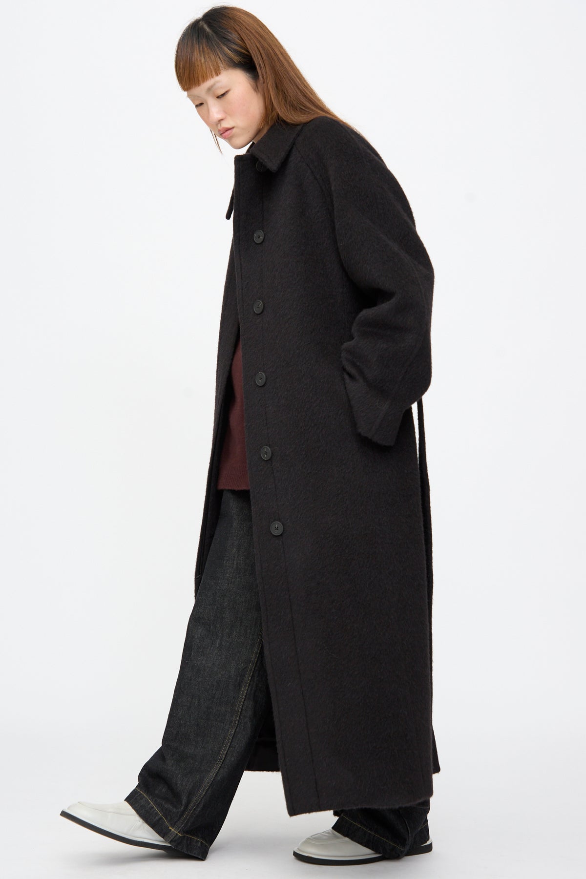 A person wearing an oversized, long dark brown textured Studio Nicholson Mohair Kober Coat in Cola over a dark top and wide-leg dark jeans with white shoes.
