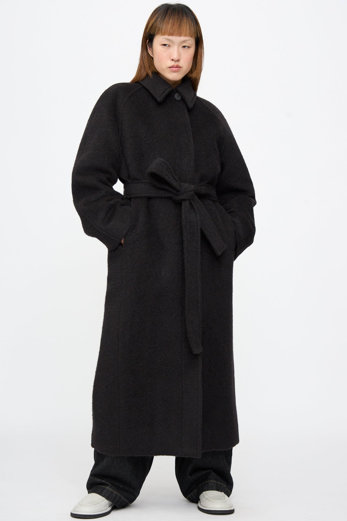 A model wears a long, oversized Studio Nicholson Mohair Kober Coat in Cola, featuring a collar and a matching belt.