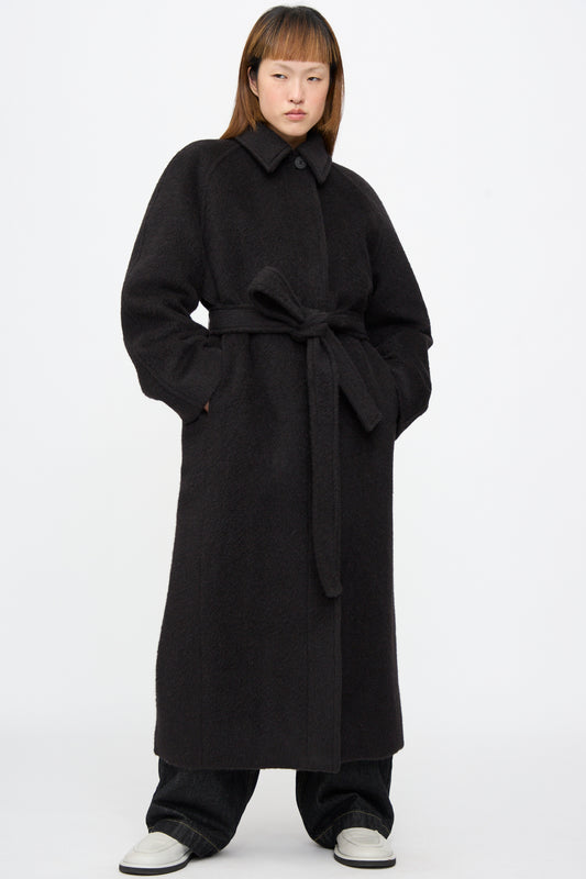 A model wears a long, oversized Studio Nicholson Mohair Kober Coat in Cola, featuring a collar and a matching belt.