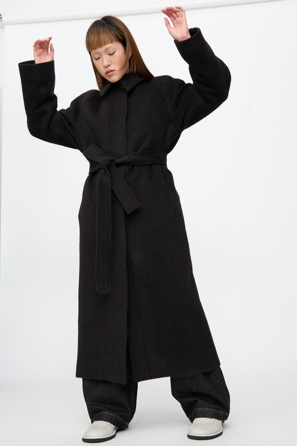 A person wearing an oversized Mohair Kober Coat in Cola, dark wide-leg trousers, and white shoes, standing with arms raised.