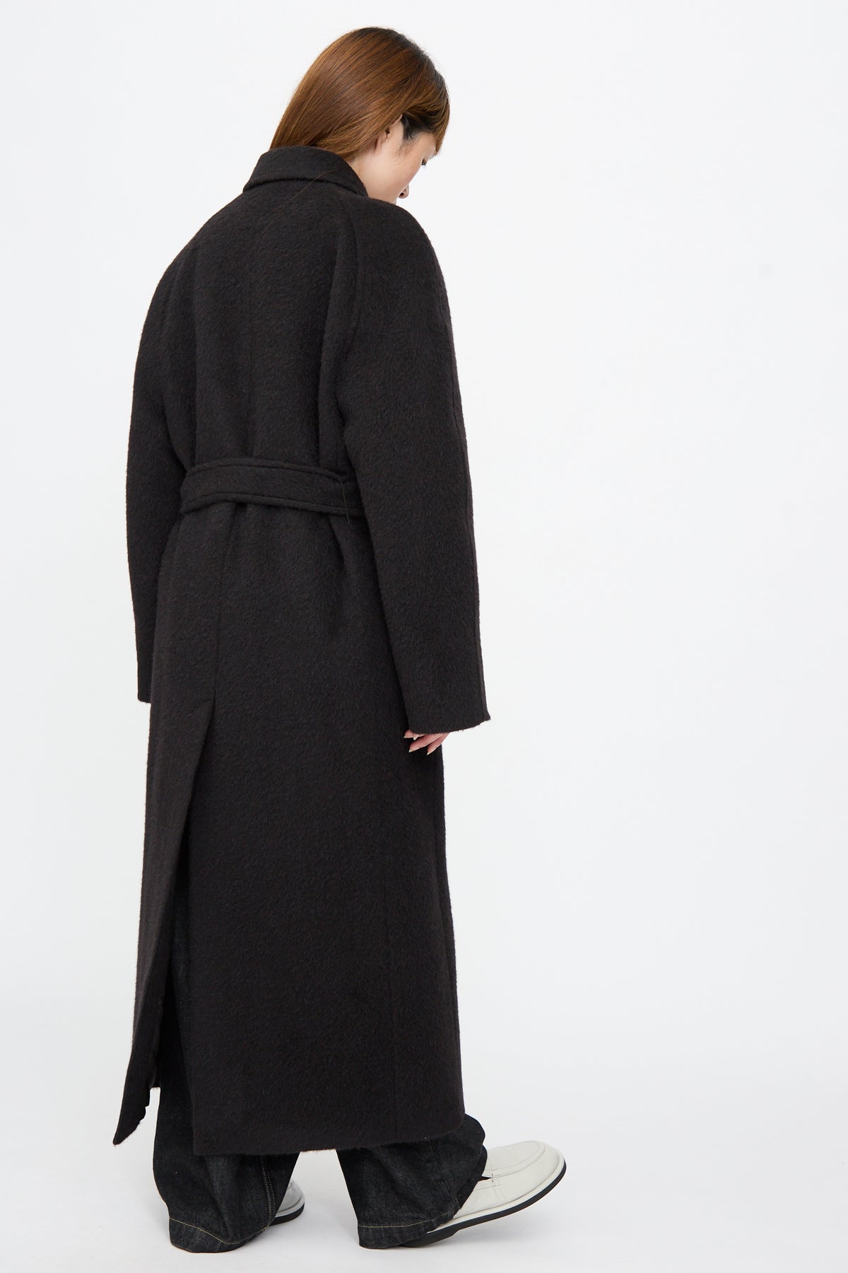 Person seen from behind wearing an oversized, dark, long Mohair Kober Coat in Cola with a belt, wide-leg trousers, and white shoes.