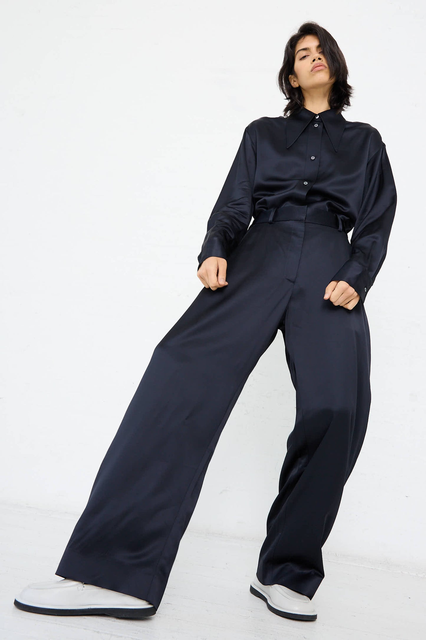 A person wearing Studio Nicholson's Myers Pant in Black Ink along with a loose-fitting, dark-colored long-sleeve shirt stands against a white background, showcasing the high-rise wide-leg design.