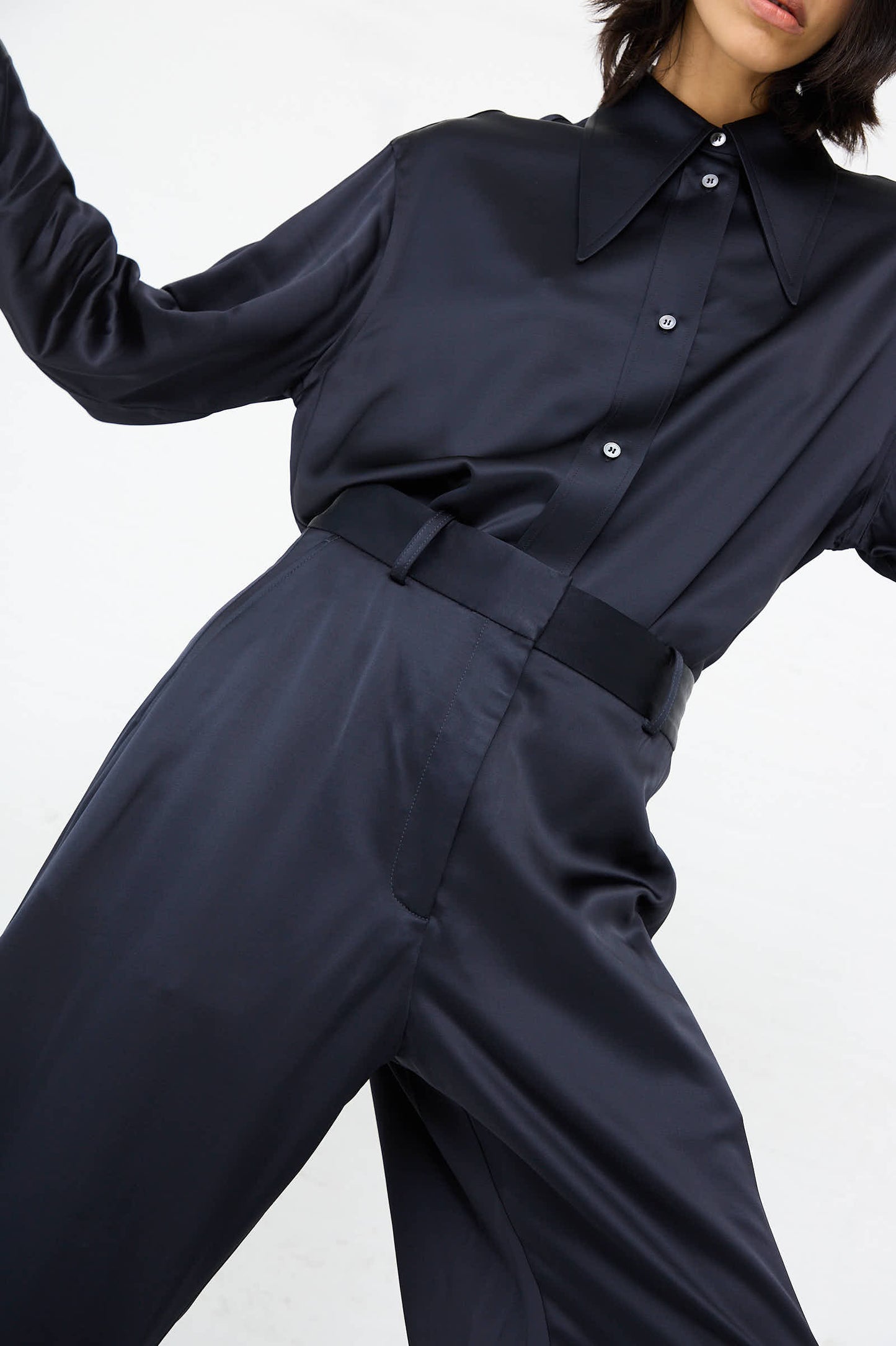 Individual clad in a dark, long-sleeve button-up shirt featuring a large pointed collar, sporting the Studio Nicholson Myers Pant in Black Ink—high-rise loose-fitting pants crafted from wool viscose satin—posing confidently against a plain white background.