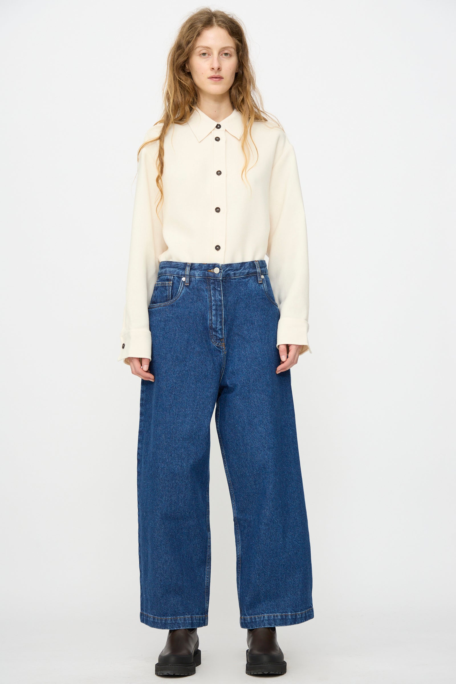 A person faces forward against a plain white background, wearing a cream button-up shirt, Studio Nicholson's Neive Curved Pant in Indigo Wash, and black boots.