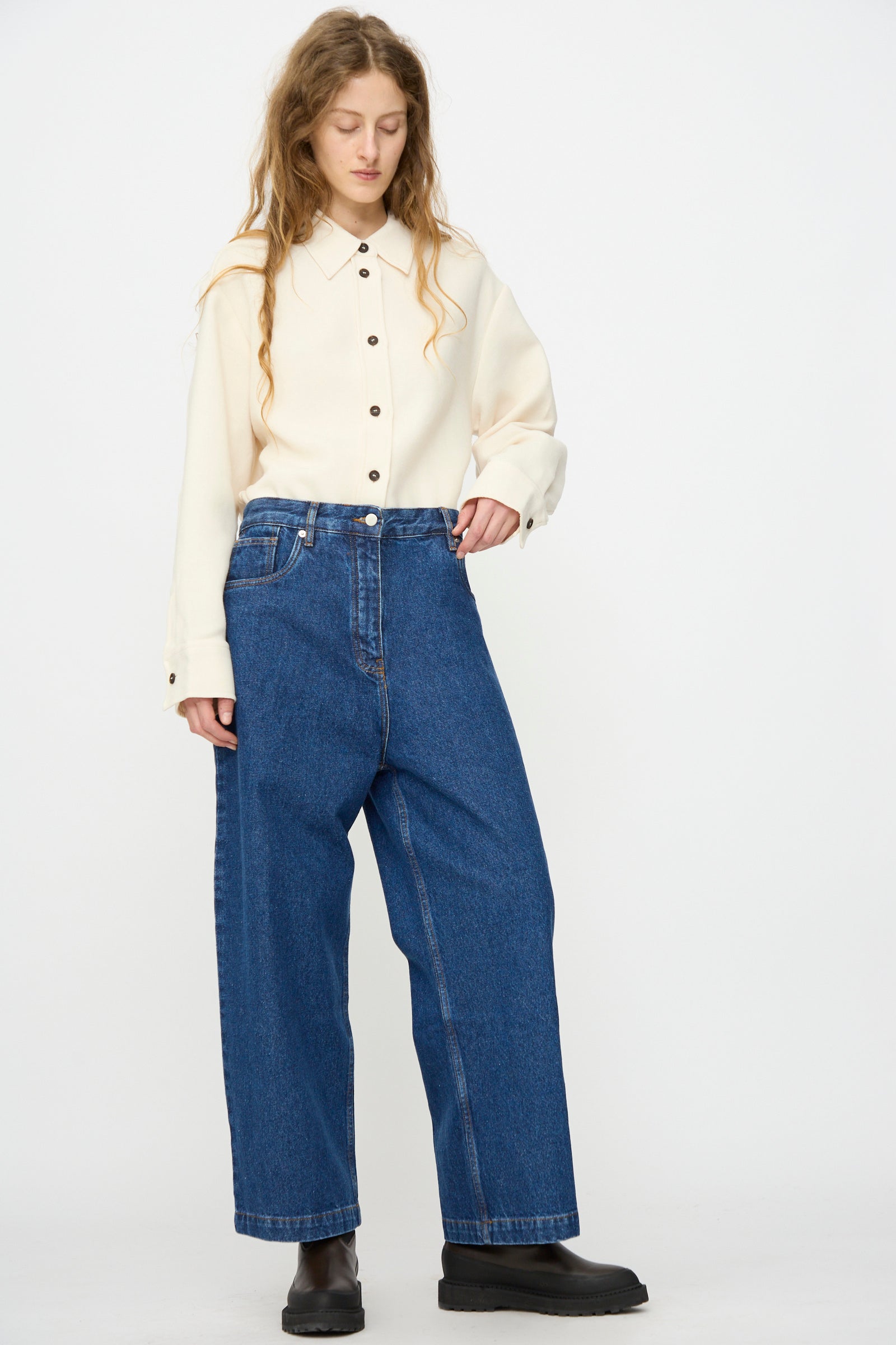 A person wearing a cream button-up shirt, Studio Nicholson Neive Curved Pant in Indigo Wash, and black shoes stands facing slightly left against a plain white background.