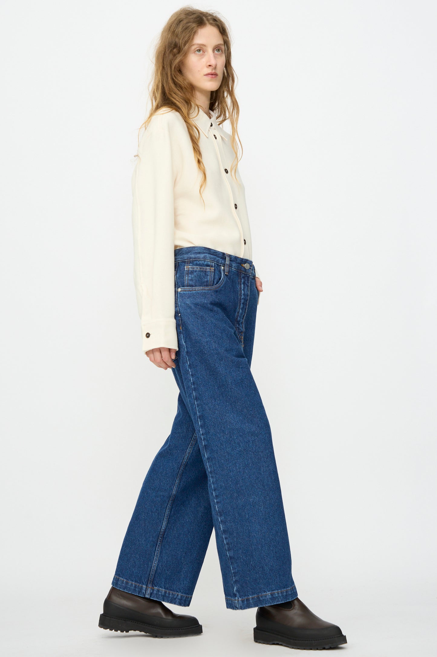 A person stands sideways against a plain white background, wearing a cream button-up shirt, black shoes, and Studio Nicholson’s Neive Curved Pant in Indigo Wash.