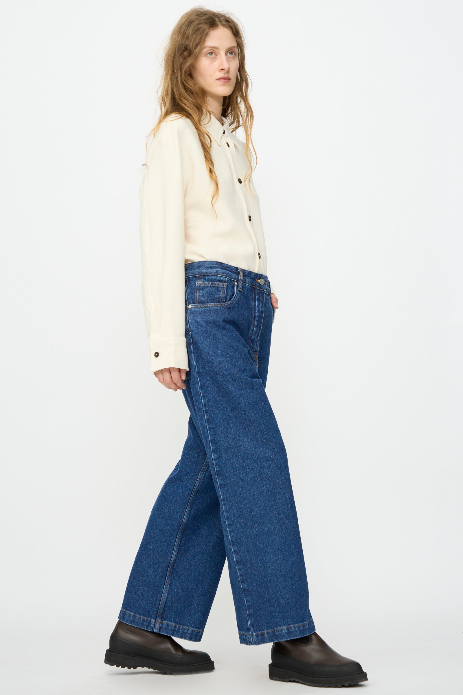 A person stands sideways against a plain white background, wearing a cream button-up shirt, black shoes, and Studio Nicholson’s Neive Curved Pant in Indigo Wash.