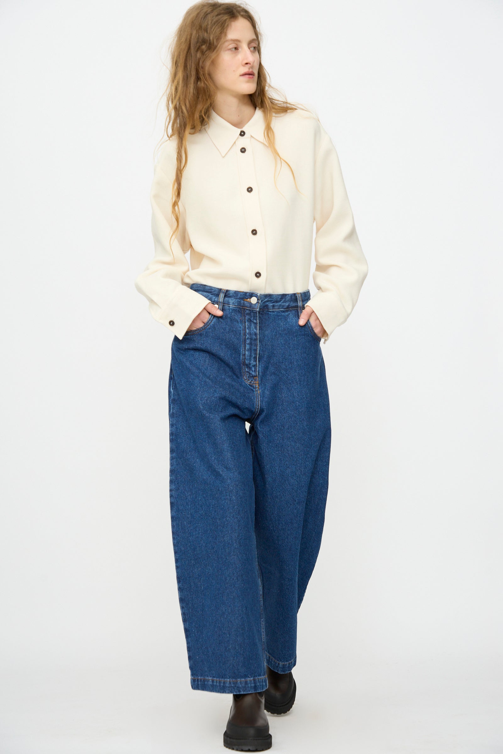 A person with long hair wears a cream button-up shirt, Studio Nicholson’s Neive Curved Pant in Indigo Wash, and black shoes, standing against a plain white background with hands in pockets.