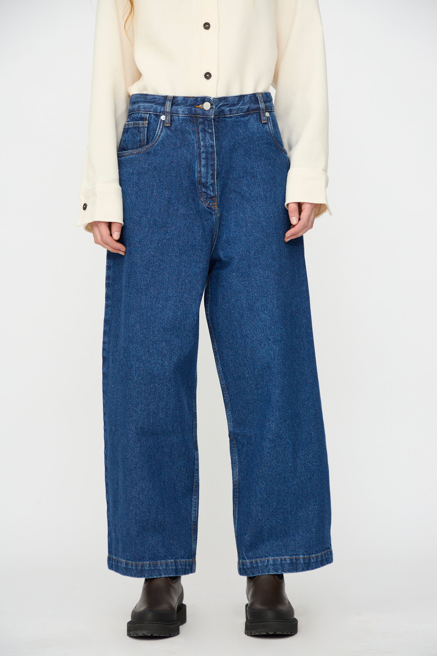 A person wears a cream button-up shirt, Studio Nicholson Neive Curved Pant in Indigo Wash, and black shoes, standing against a plain background.