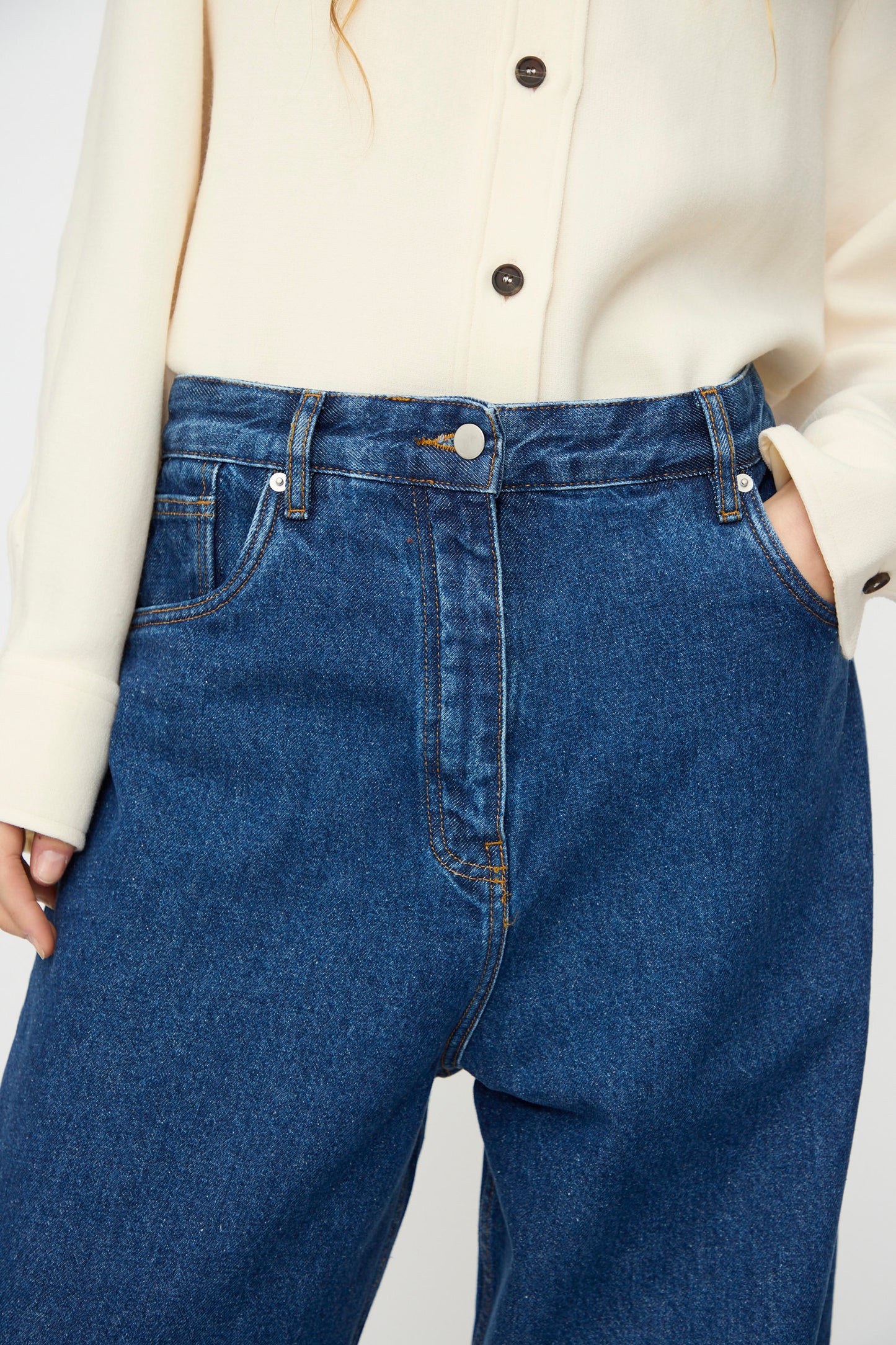 Wearing a cream button-up shirt tucked into Studio Nicholson’s Neive Curved Pant in Indigo Wash, with hands in the pockets.