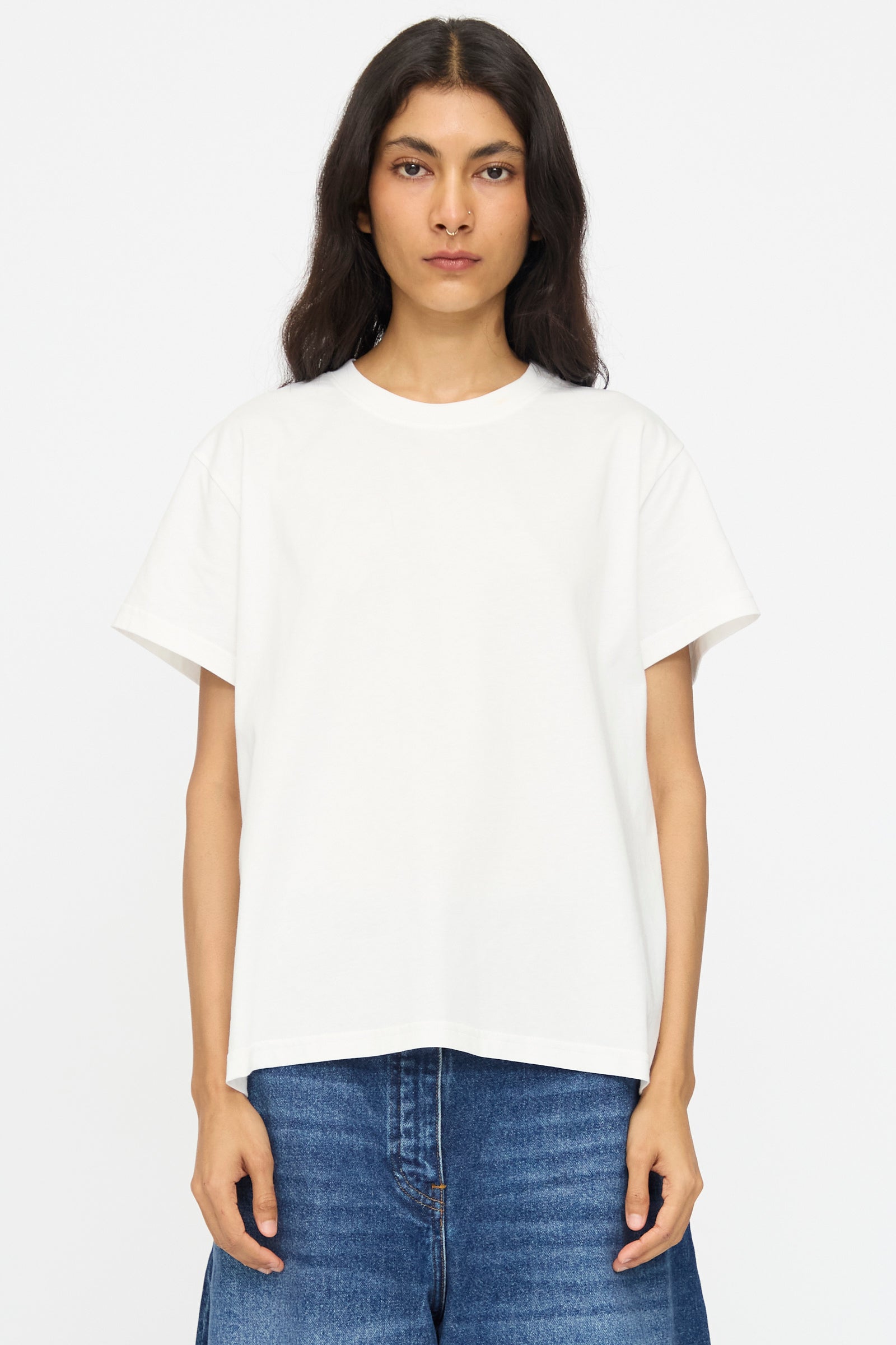 A person with long dark hair wears the Studio Nicholson Perfect Marine Tee in Off White and blue jeans, standing against a white background.