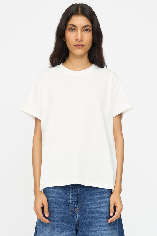 A person with long dark hair wears the Studio Nicholson Perfect Marine Tee in Off White and blue jeans, standing against a white background.