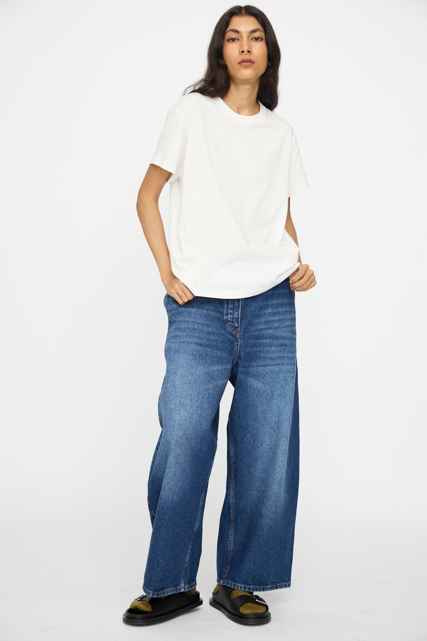 A person stands against a plain background wearing the Studio Nicholson Perfect Marine Tee in Off White, styled with wide-leg blue jeans and black sandals paired with yellow socks.