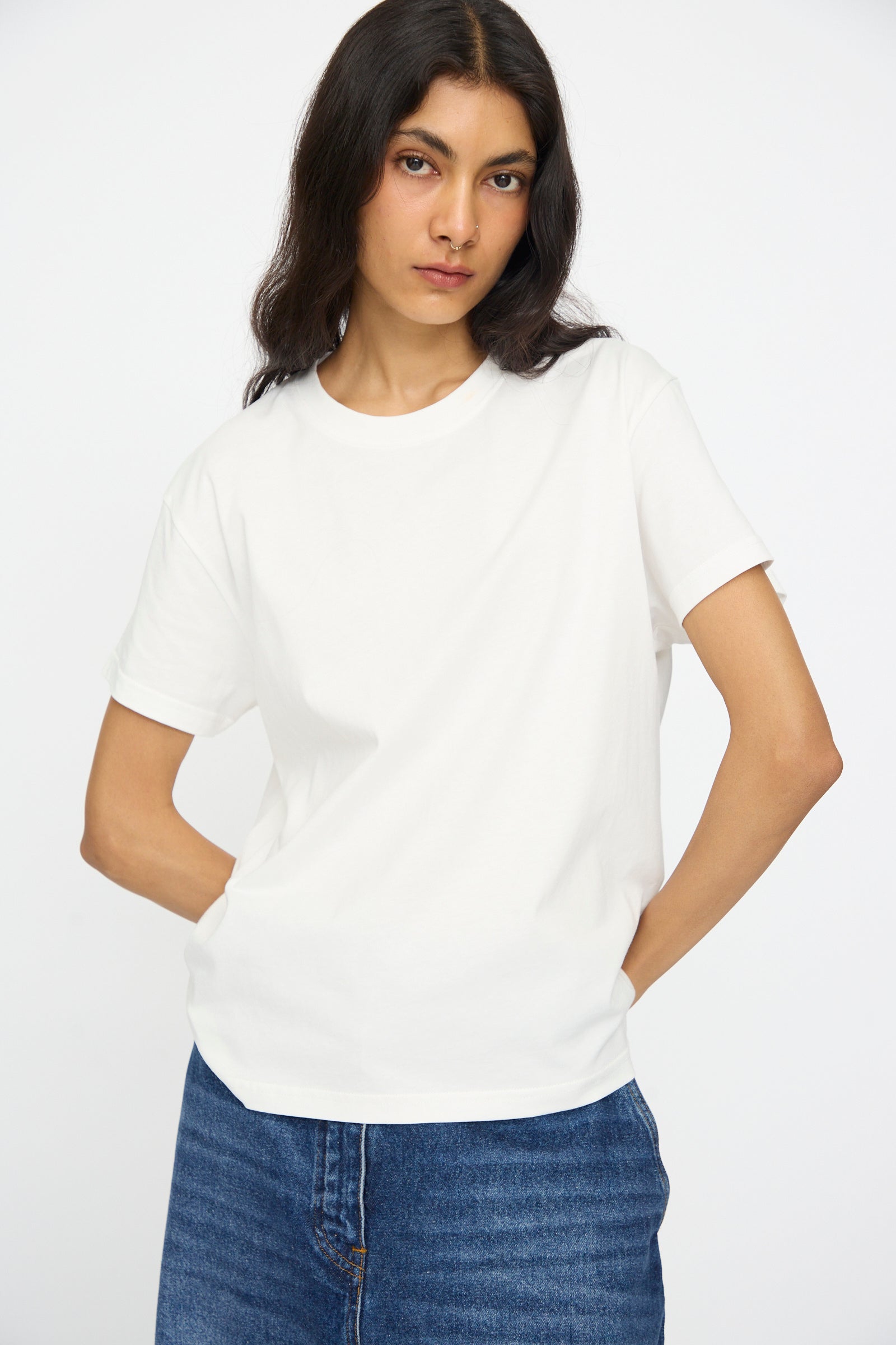 A person with long dark hair wears the Studio Nicholson Perfect Marine Tee in Off White and blue jeans, standing with hands in pockets against a plain light background.