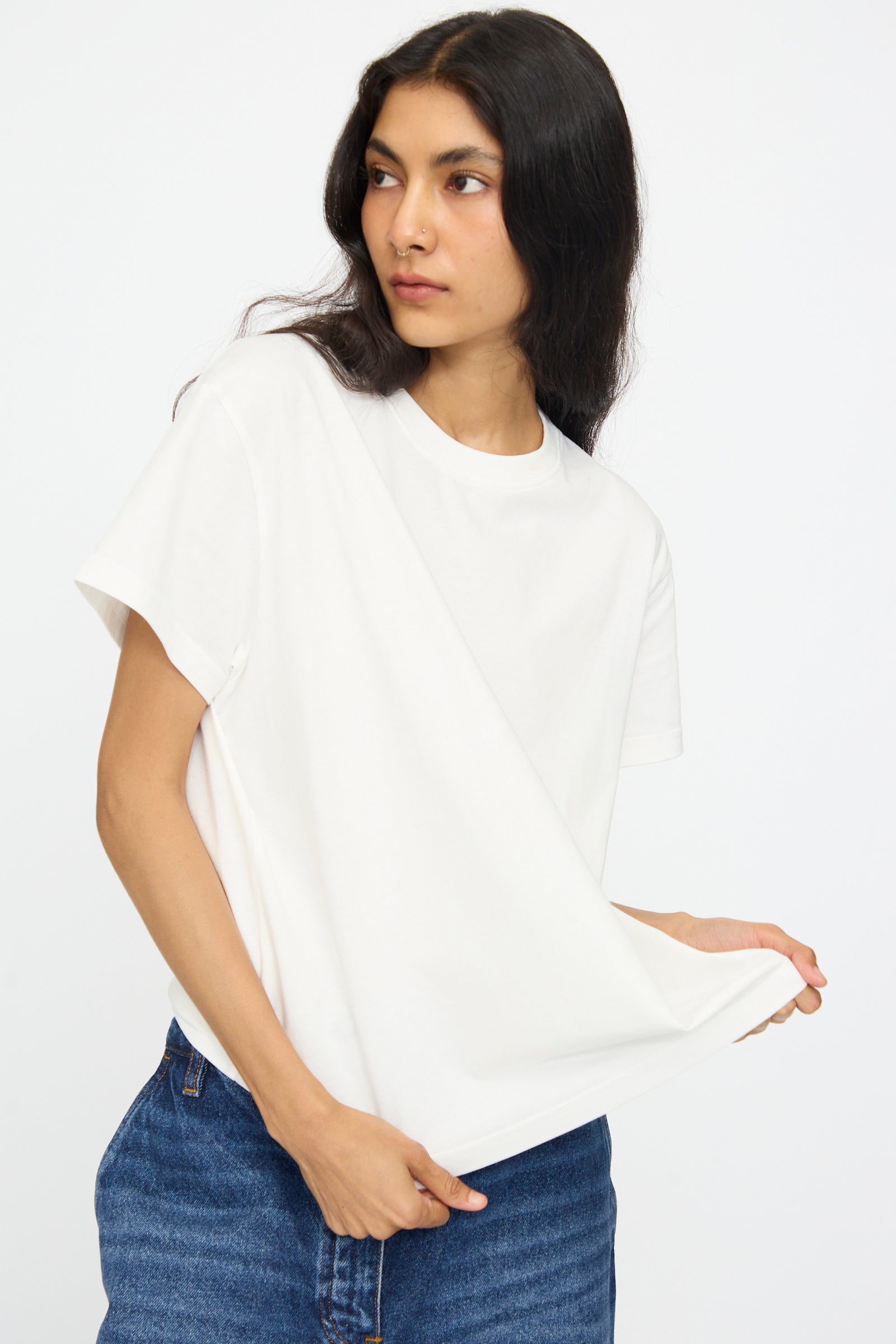 A person wears the Studio Nicholson Perfect Marine Tee in Off White, styled with blue jeans, standing against a white background and holding out the shirt hem while looking to the side.