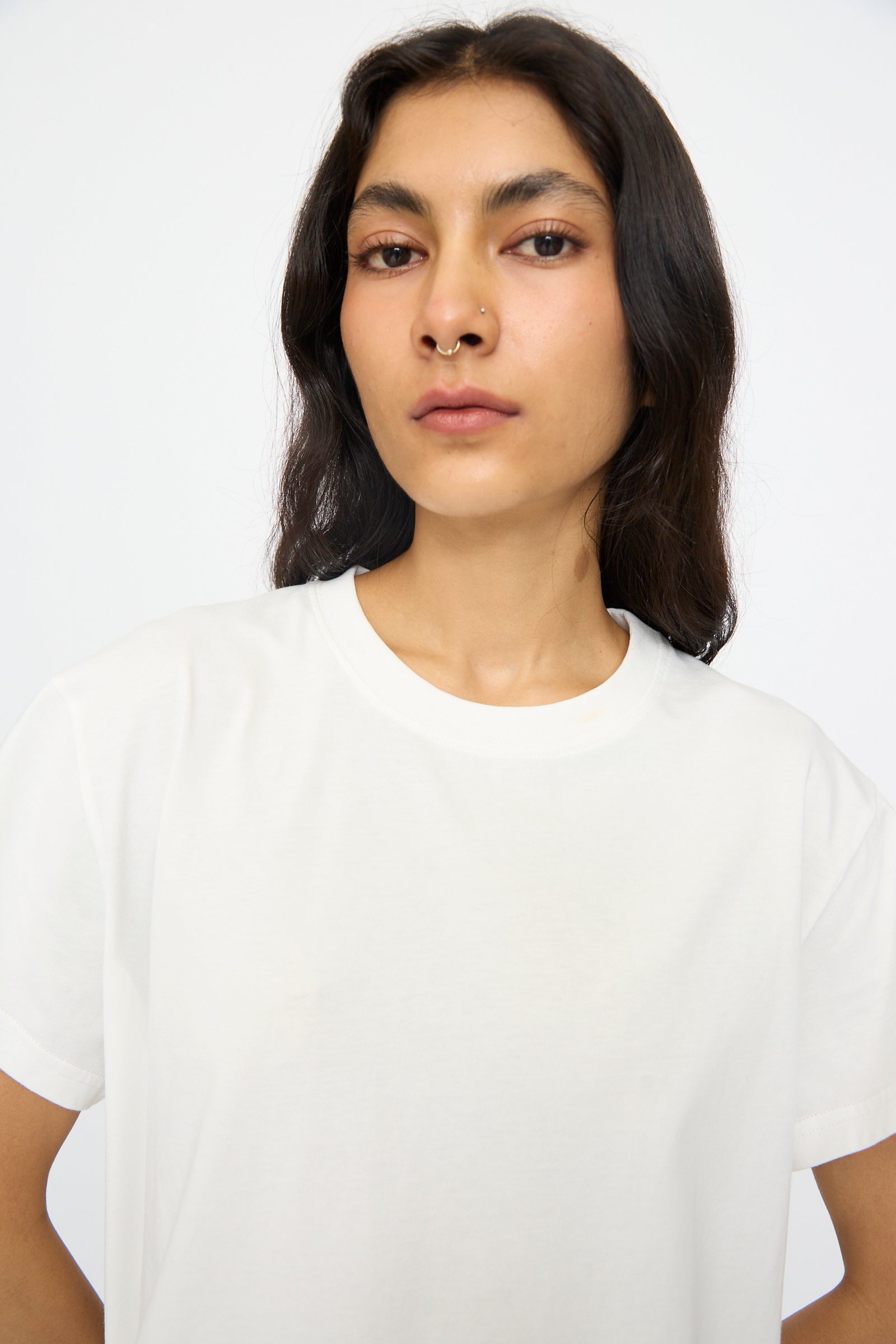 A person with long dark hair and a nose ring wears the Studio Nicholson Perfect Marine Tee in Off White, standing against a white background.