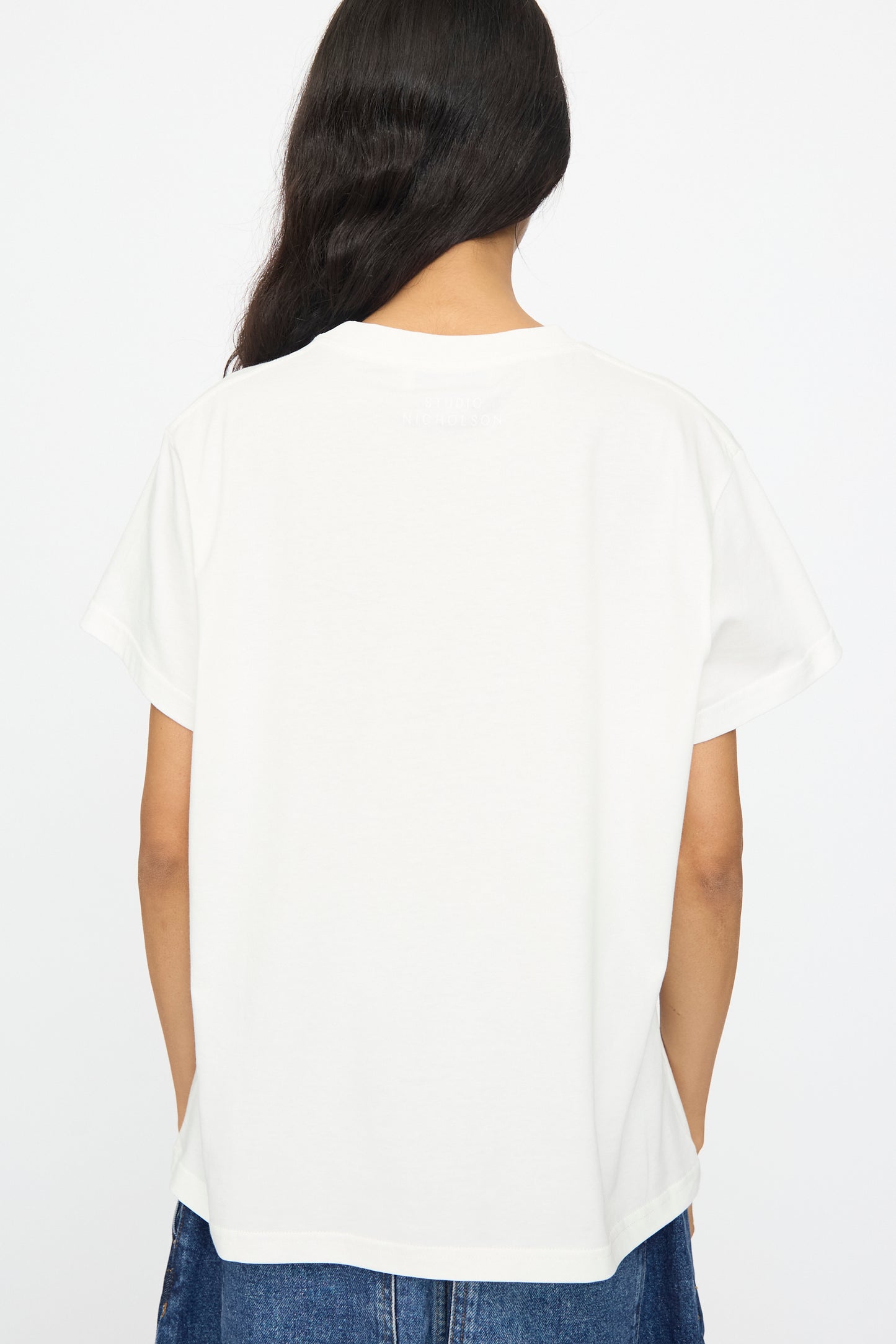 Person with long dark hair shown from the back wearing Studio Nicholson’s Perfect Marine Tee in Off White, featuring a boxy fit and short sleeves, paired with blue jeans against a plain white background.
