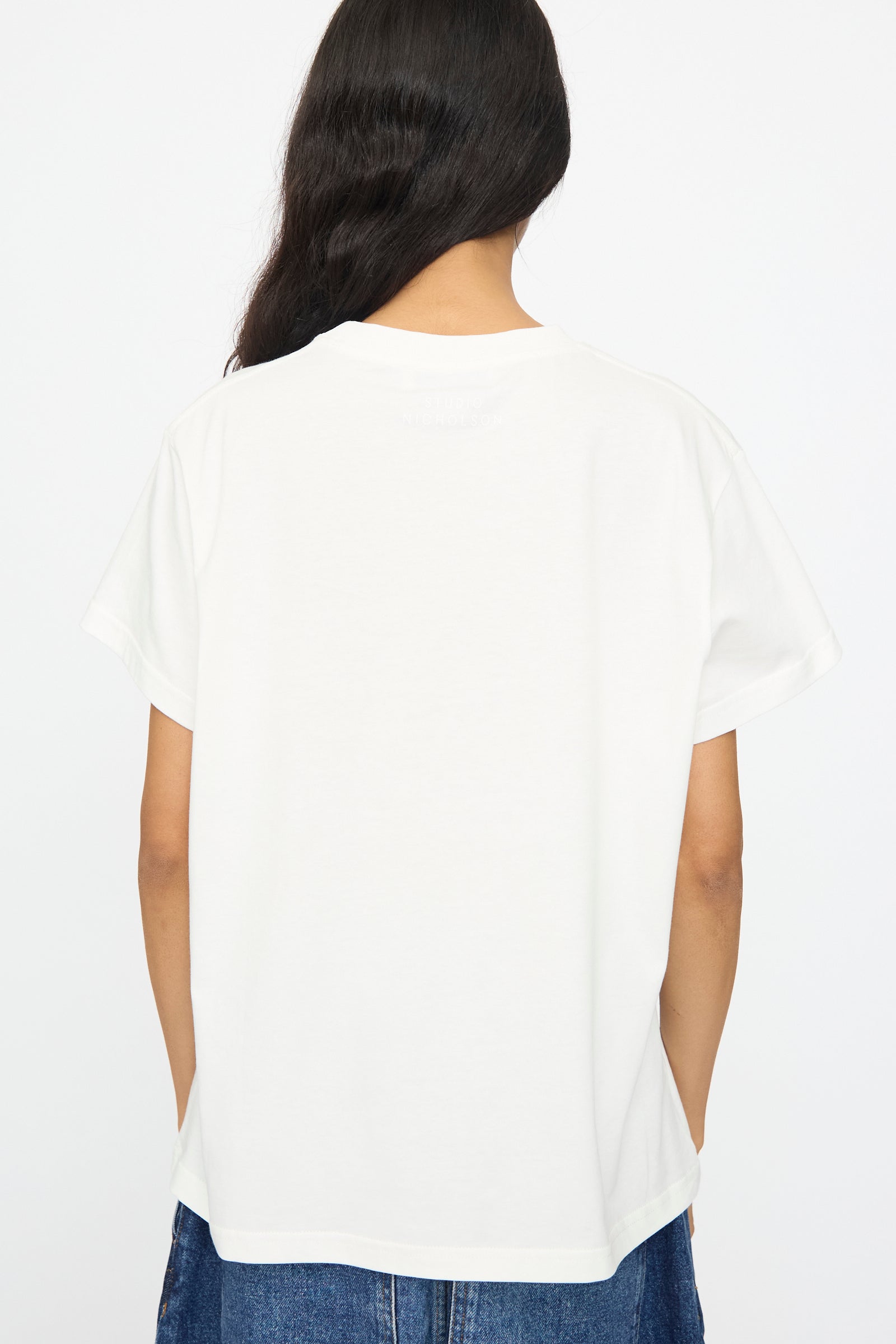 Person with long dark hair shown from the back wearing Studio Nicholson’s Perfect Marine Tee in Off White, featuring a boxy fit and short sleeves, paired with blue jeans against a plain white background.