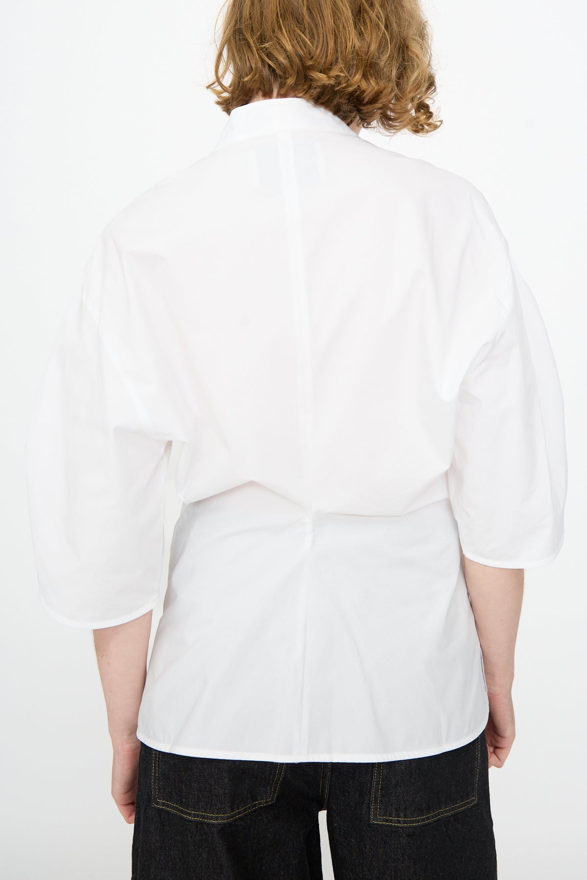 A person wearing the Studio Nicholson Powder Cotton Asti Top in White—a loose-fitting, three-quarter sleeve shirt with a fitted waist—paired with black pants, seen from the back against a plain white background.