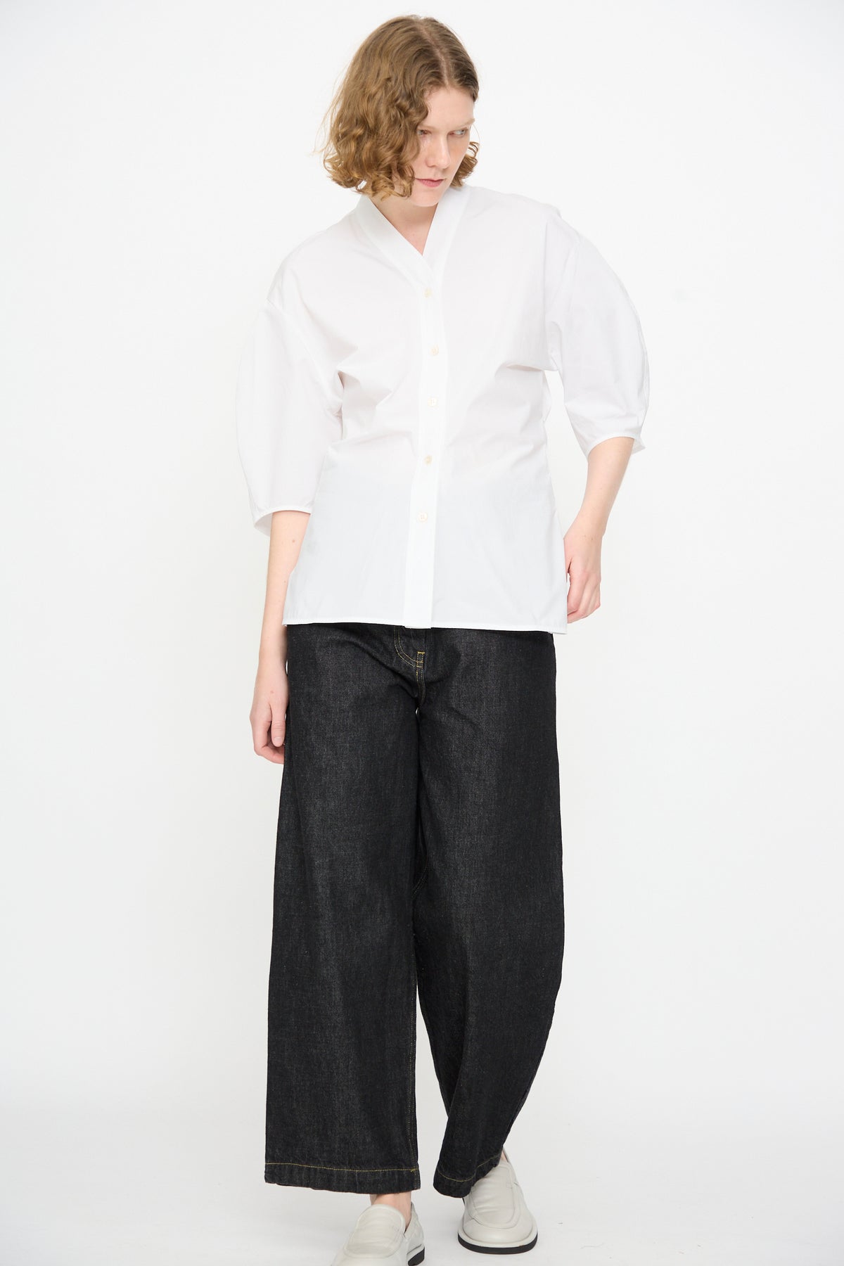 A person stands against a white background, wearing the Studio Nicholson Powder Cotton Asti Top in White—a loose button-up shirt with a V-neckline—paired with wide-leg black jeans and white shoes, with one hand at their side and the other on their hip.