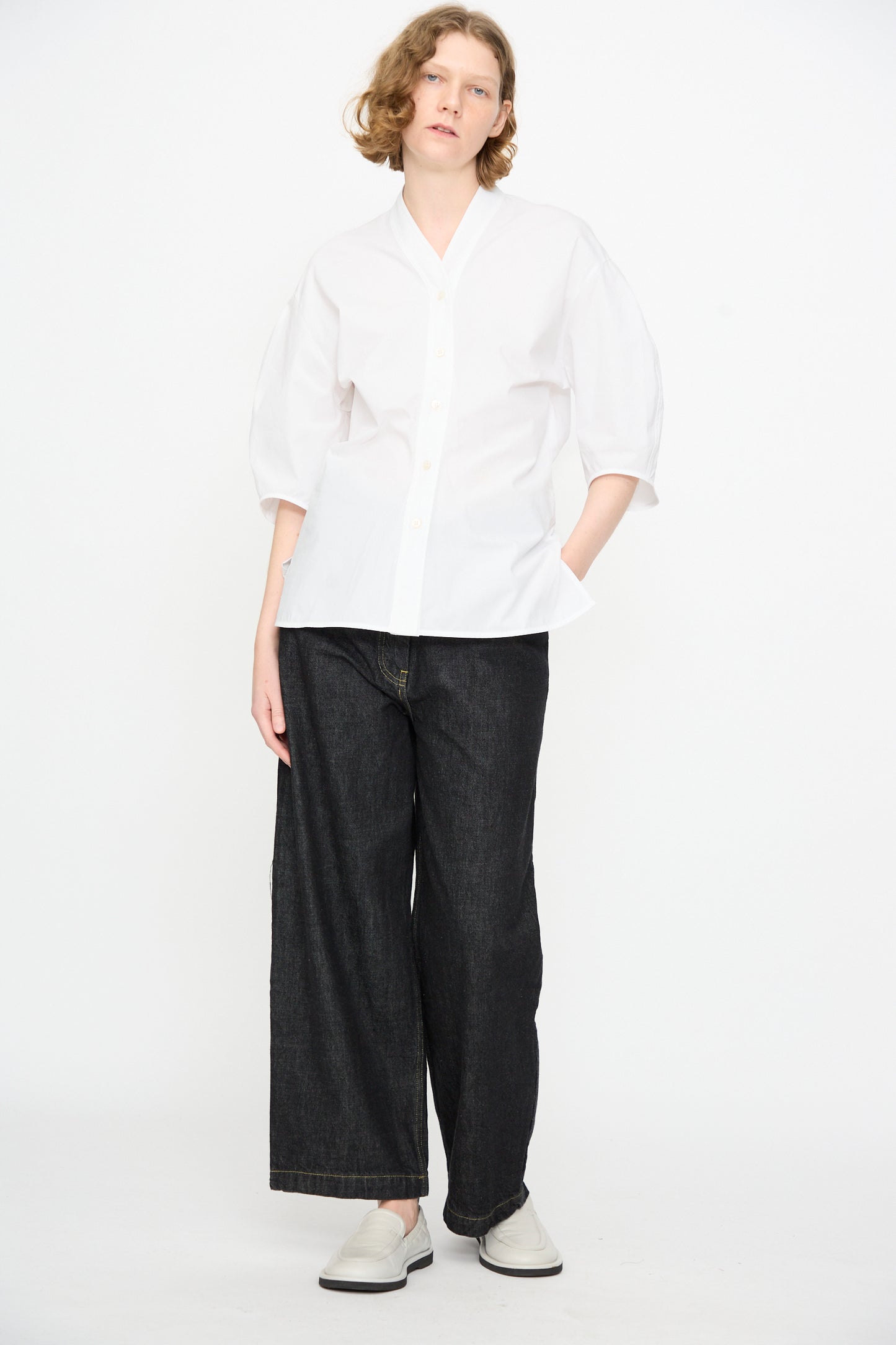 A person stands against a white background, wearing the Studio Nicholson Powder Cotton Asti Top in White—a loose button-up shirt with a subtle V-neckline—paired with wide-leg dark jeans and white shoes.