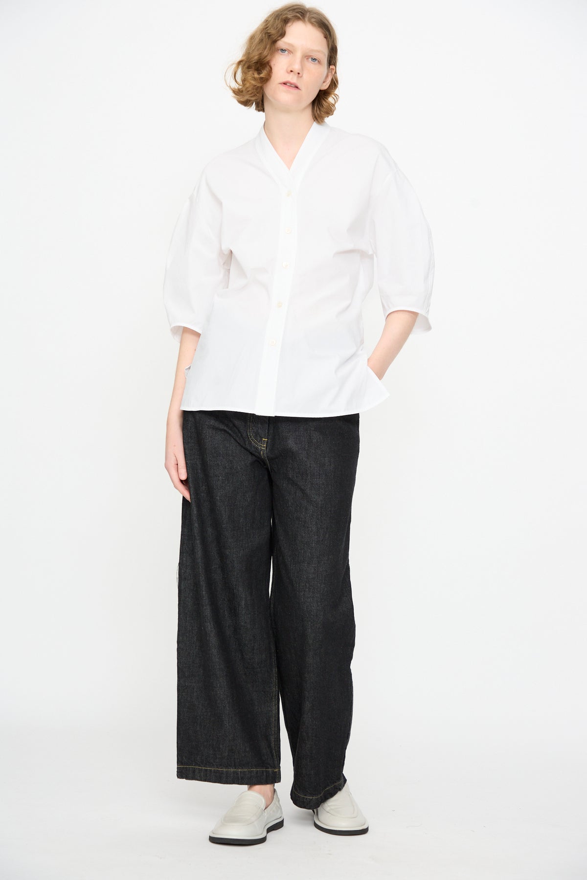 A person stands against a white background, wearing the Studio Nicholson Powder Cotton Asti Top in White—a loose button-up shirt with a subtle V-neckline—paired with wide-leg dark jeans and white shoes.