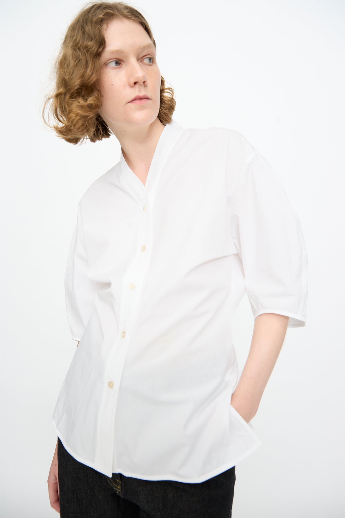 A person with short, wavy light brown hair wears the Studio Nicholson Powder Cotton Asti Top in White—a loose button-up shirt with a subtle V-neckline—paired with dark pants, standing against a plain white background and facing slightly to the side.