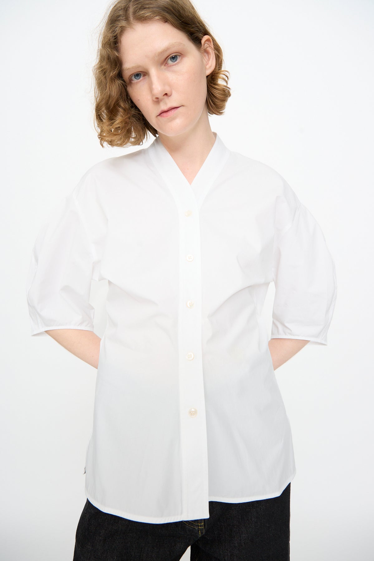 A person wearing the Studio Nicholson Powder Cotton Asti Top in White—a loose-fitting button-up shirt featuring three-quarter sleeves and a subtle V-neckline—stands against a plain white background with their hands in their pockets.