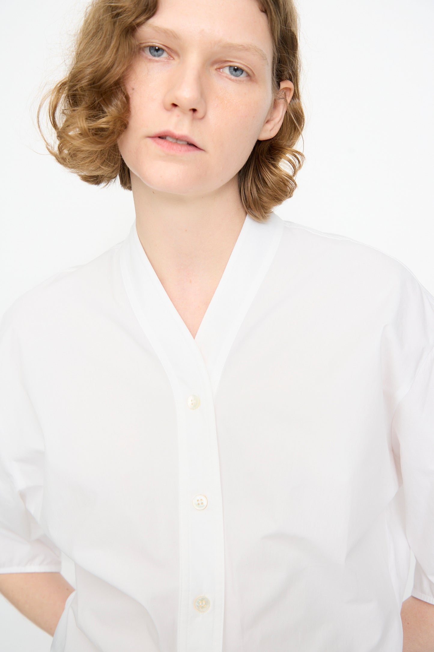 A person with short, wavy light brown hair is wearing the Studio Nicholson Powder Cotton Asti Top in White—a white button-up shirt featuring a V-neckline—standing against a plain white background and looking directly at the camera.