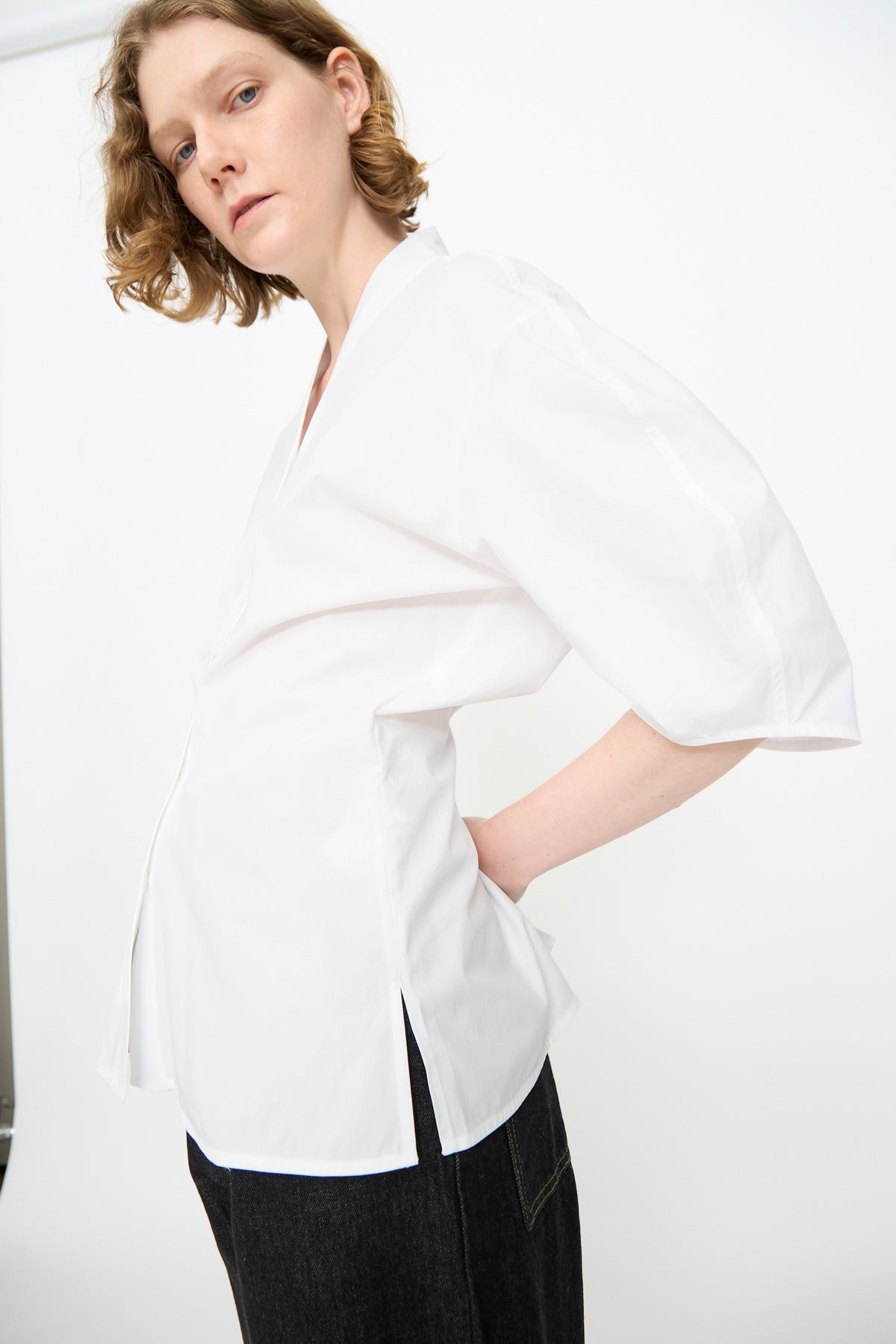 A person with short, wavy hair stands in profile against a plain white background, wearing the Studio Nicholson Powder Cotton Asti Top in White—a loose-fitting shirt with a V-neckline—paired with dark pants.