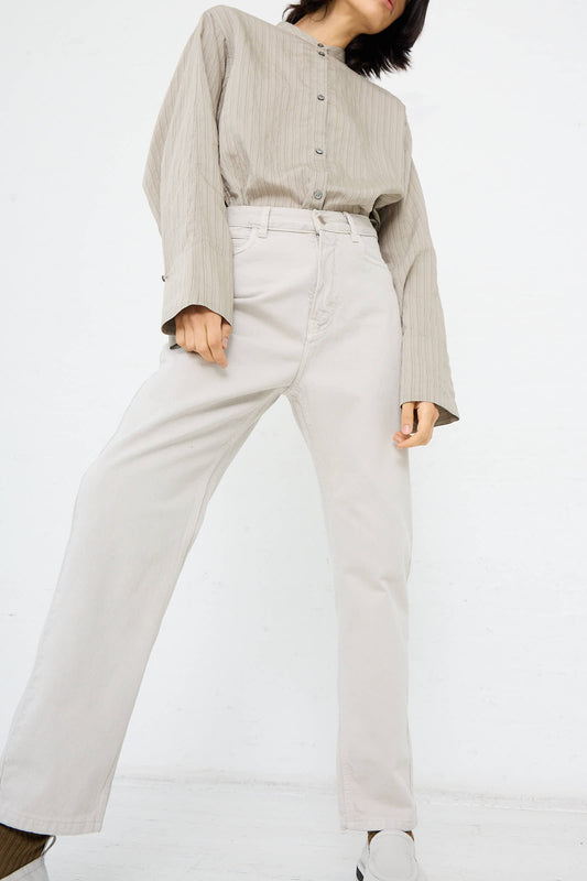 A person wearing a loose-fitting, button-up shirt and the high-waisted Studio Nicholson Ruthe Boyfriend Jean in Bone Grey stands with legs apart against a plain white background.