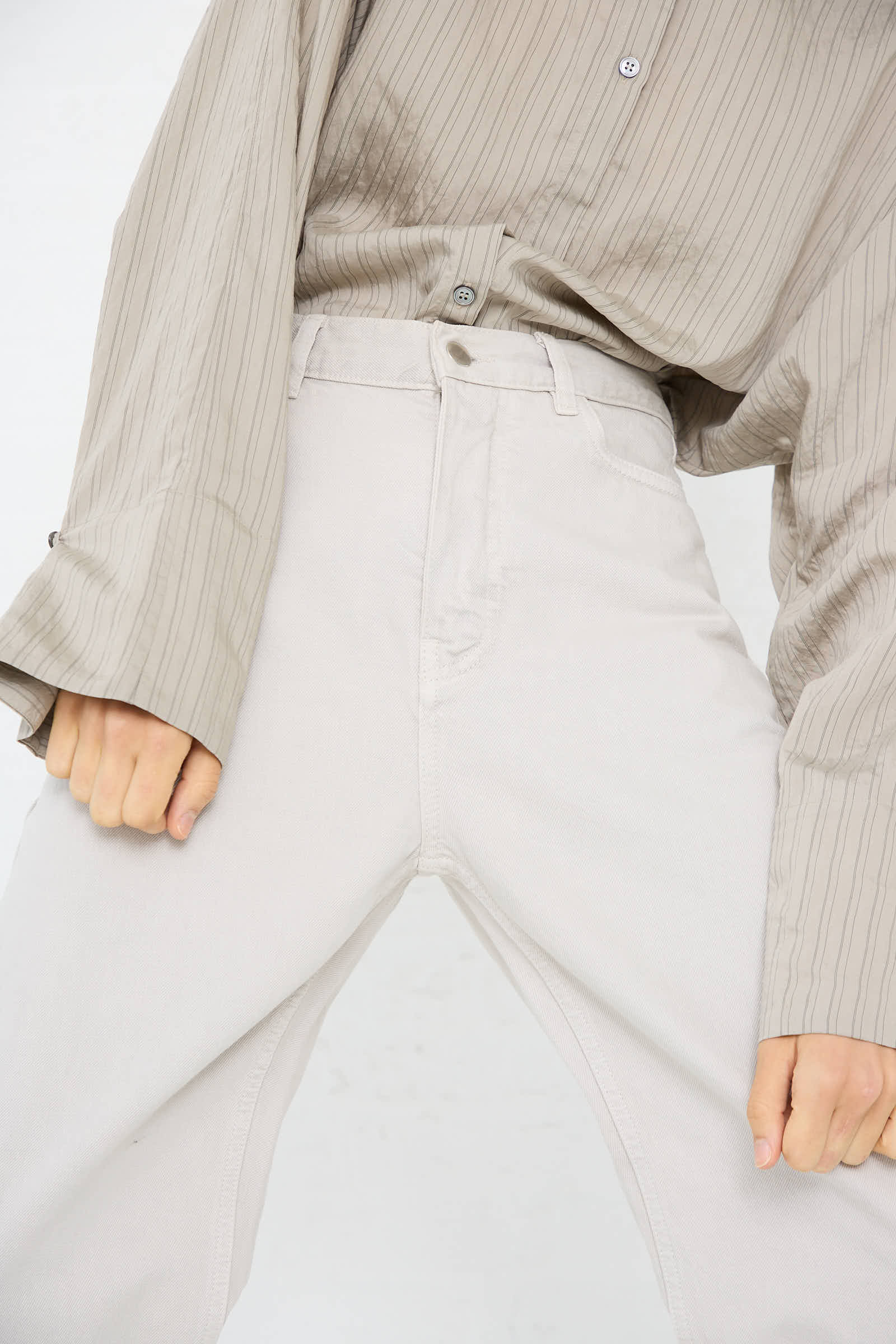 A person wearing a loose, striped, taupe-colored shirt and the Ruthe Boyfriend Jean in Bone Grey by Studio Nicholson, with a high waist, partially bending forward. The image focuses on the torso and legs.