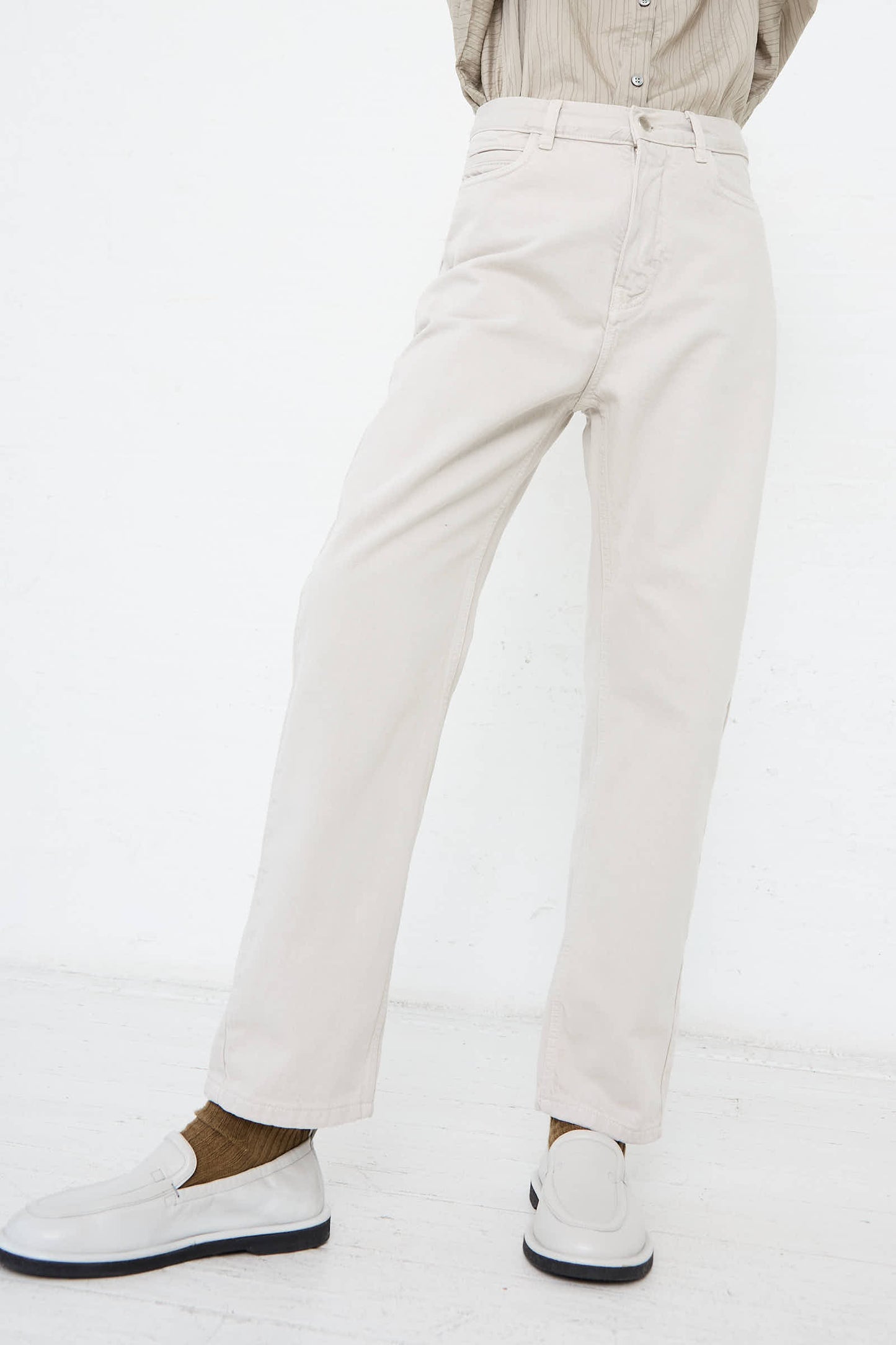 A person sporting the Ruthe Boyfriend Jean in Bone Grey from Studio Nicholson, paired with a beige high-rise trouser, a beige shirt, and white loafers, stands against a plain white background, capturing an elegant yet classic style.