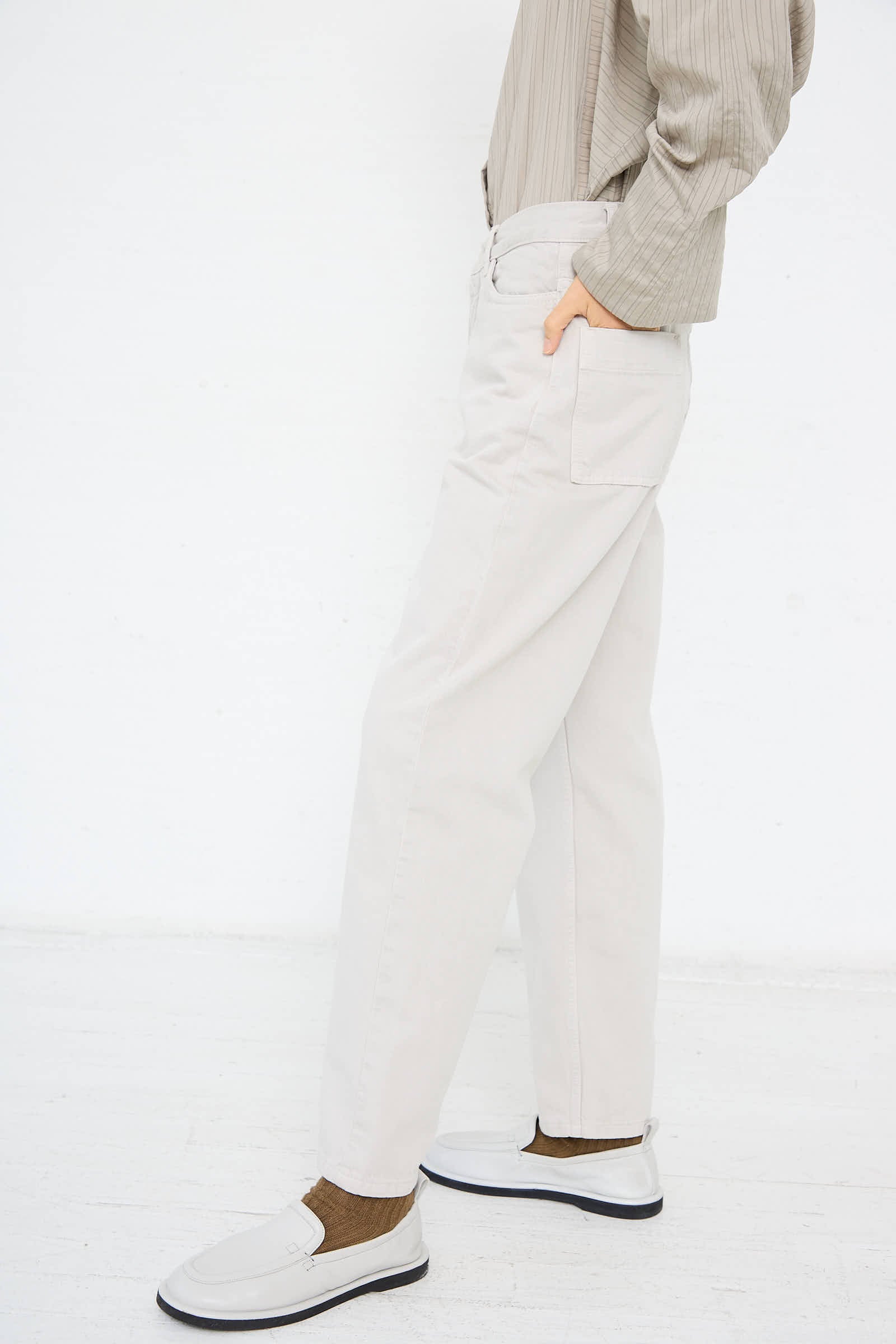 A person wearing Studio Nicholson's Ruthe Boyfriend Jean in Bone Grey, a striped beige shirt, and white slip-on shoes stands against a white background.