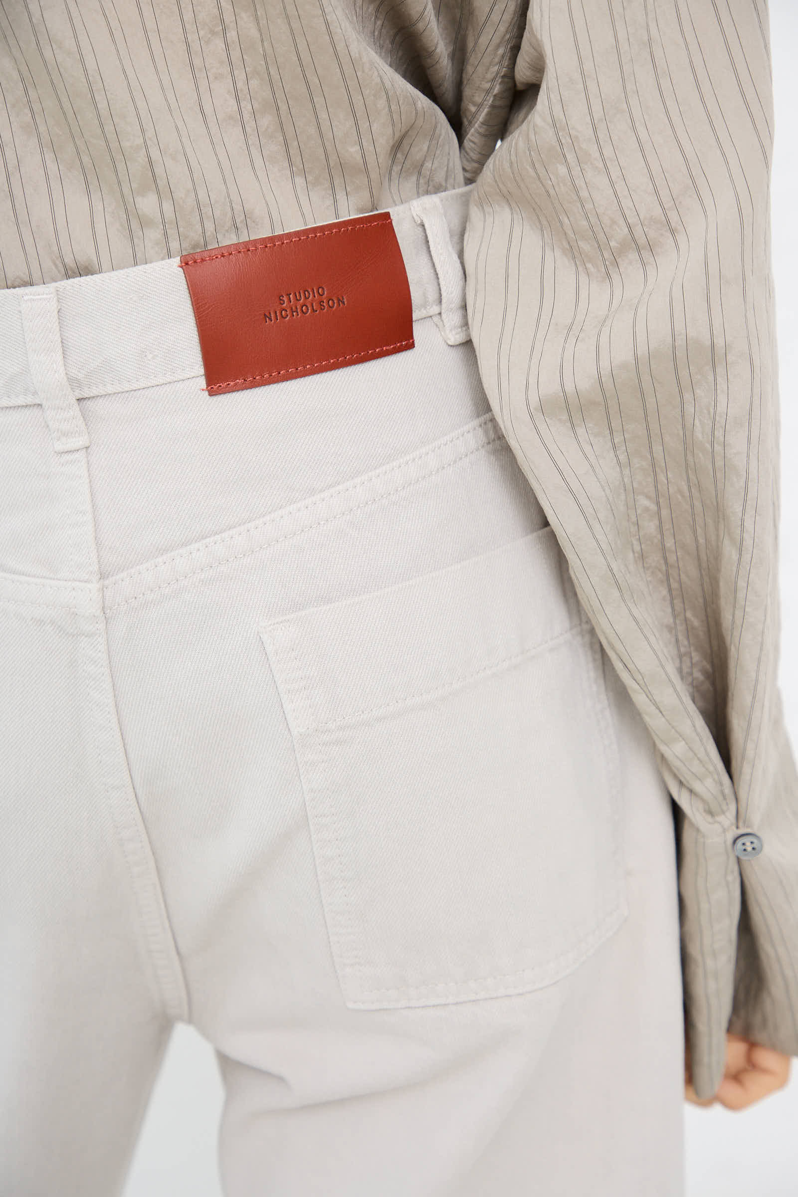 Close-up of a person wearing Studio Nicholson's light-colored Ruthe Boyfriend Jean in Bone Grey with a visible back pocket and a brown Studio Nicholson label, paired with a beige pinstripe shirt.