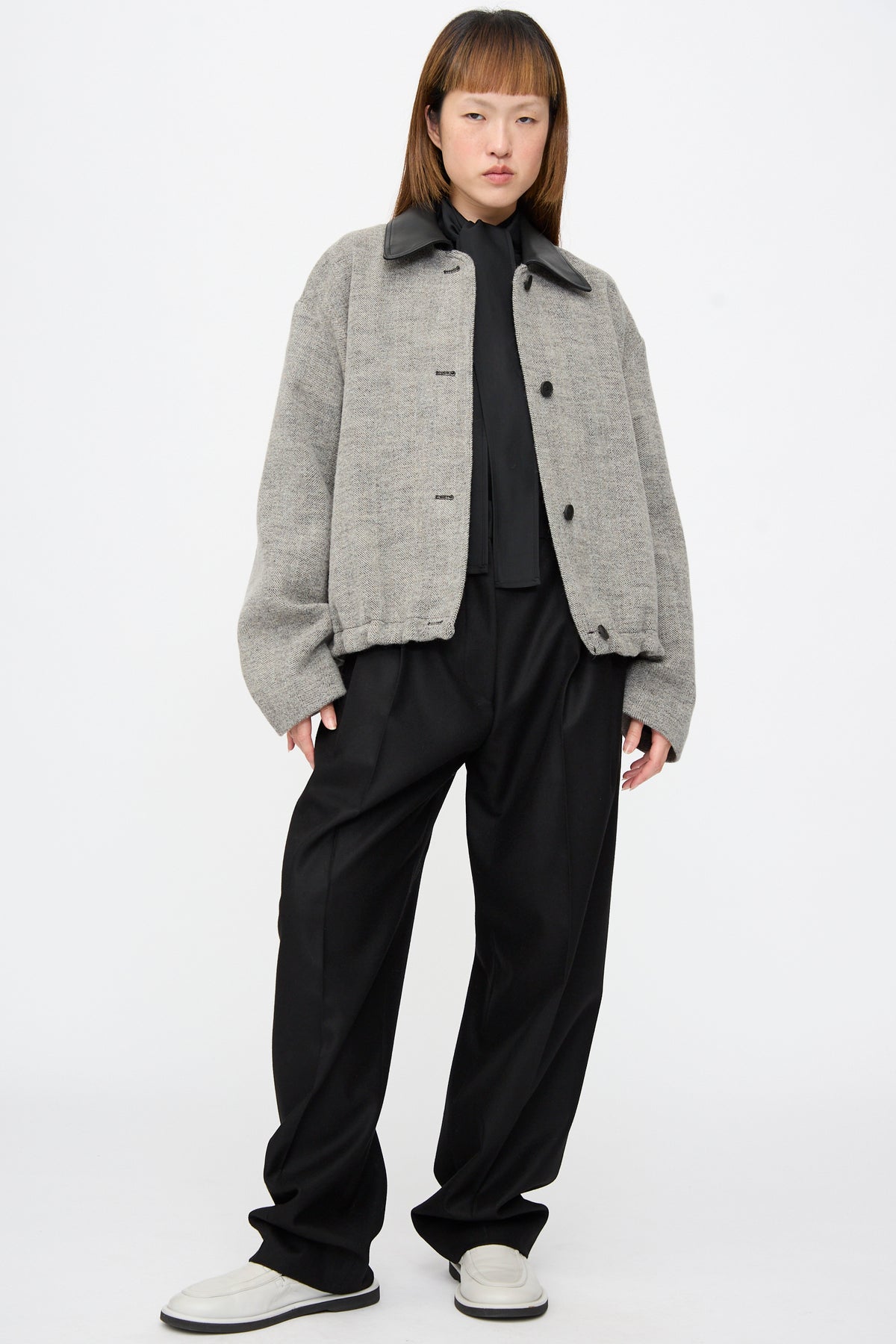 A person wearing a grey herringbone Studio Nicholson Seberg Chore Jacket in Black and White with a black collar over a black shirt and black trousers.