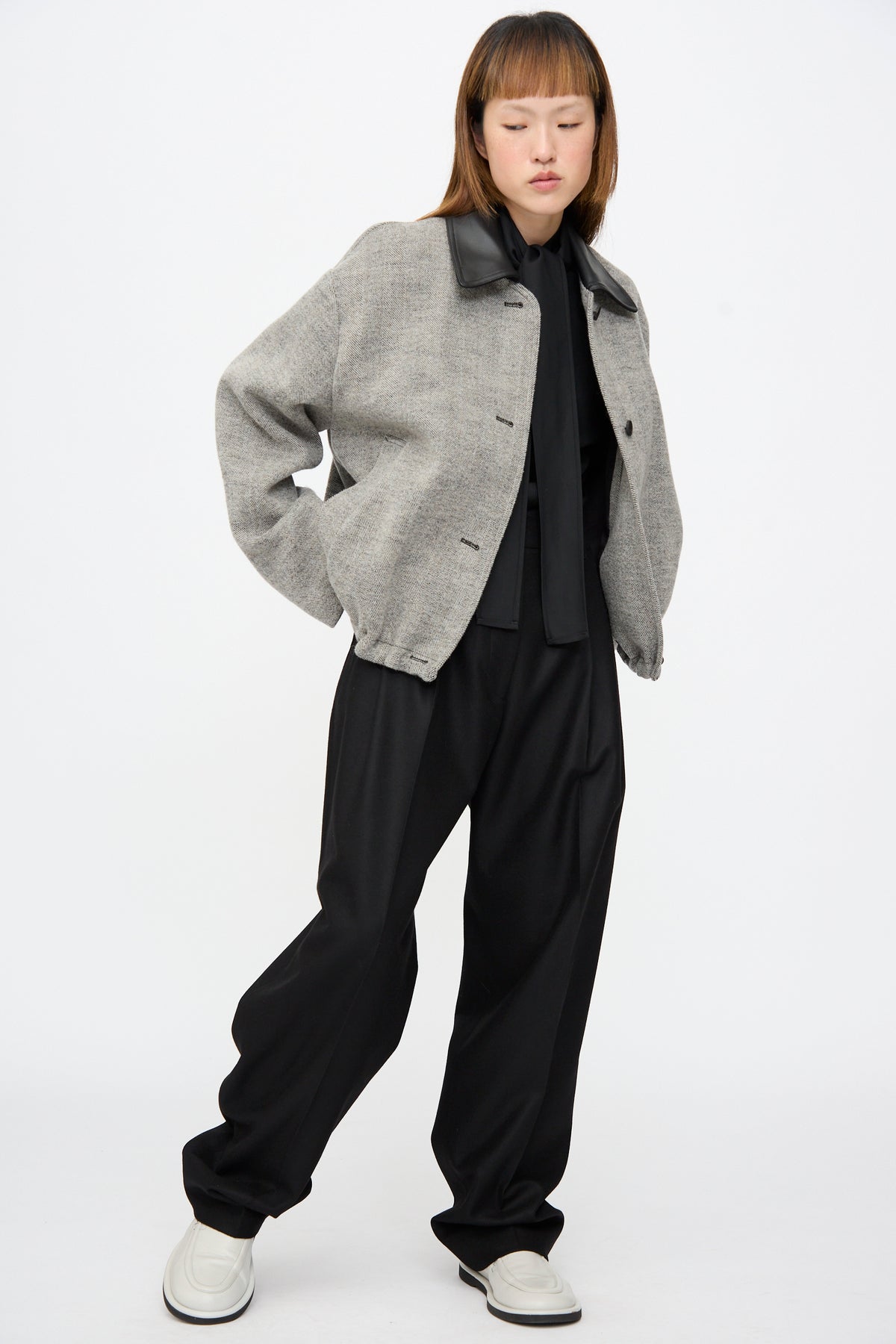 A person wears a Studio Nicholson Seberg Chore Jacket in Black and White over a black shirt and black trousers, paired with white loafers.