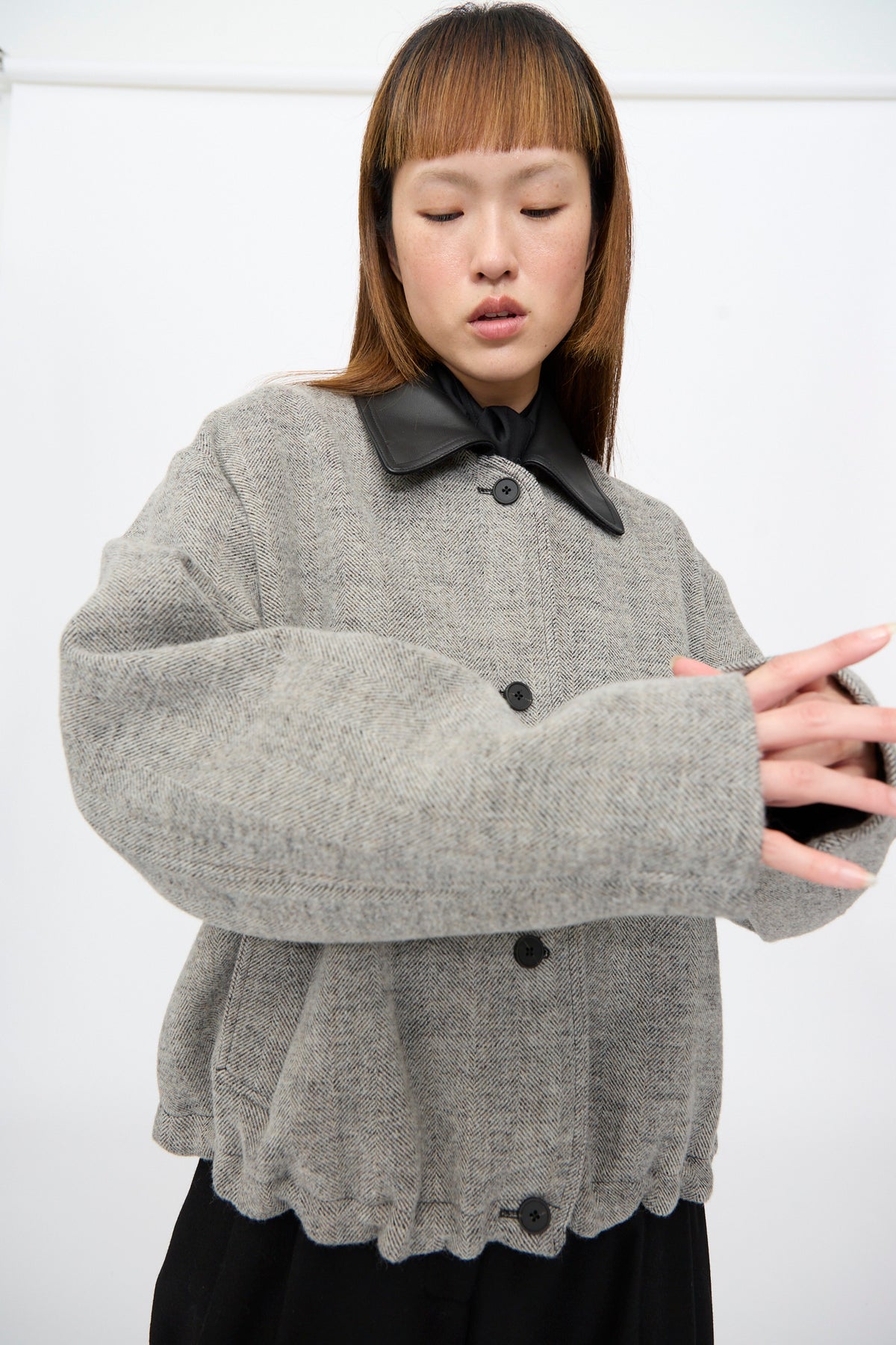 A person wearing a Studio Nicholson Seberg Chore Jacket in Black and White, featuring a grey herringbone pattern, leather collar, and a cinched, elasticated hem.